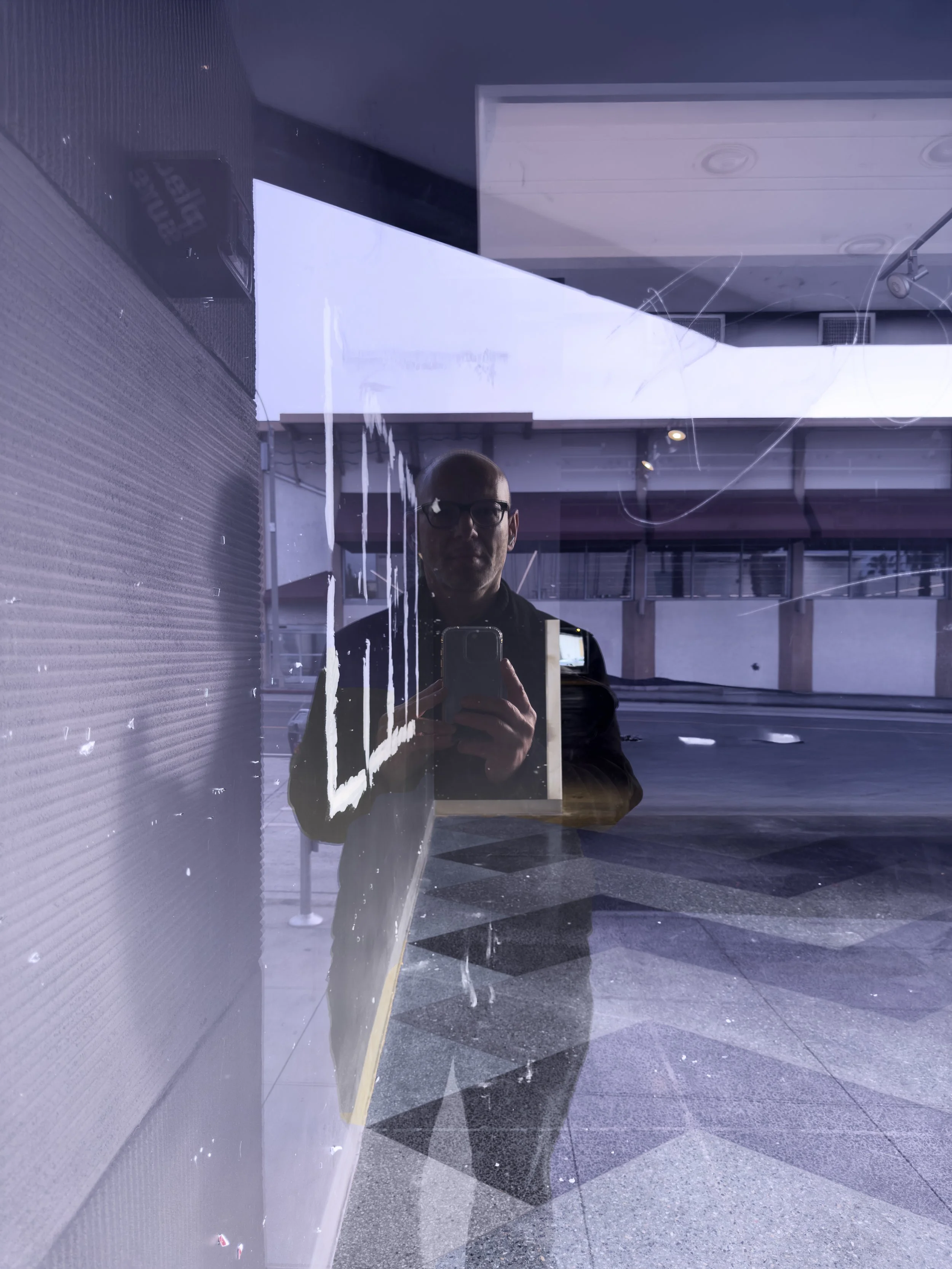 Reflection of a man taking a photo with a phone in a glass window or door, with visible scratches or markings on the glass.