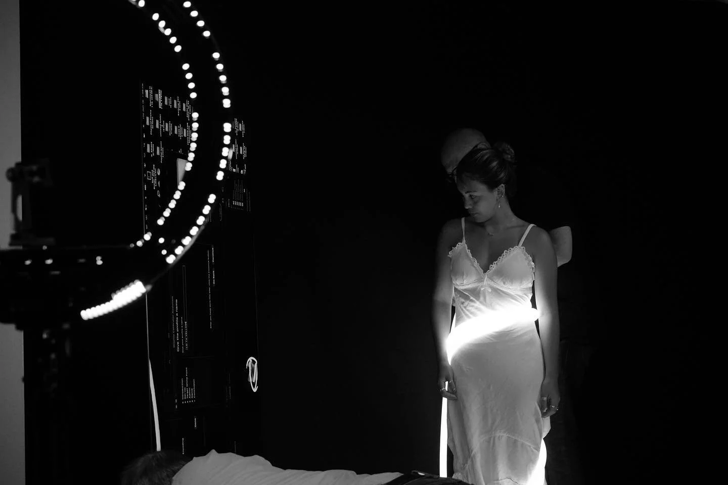 Black and white photo of a woman in a white dress and a man behind her in a dim setting, illuminated by a ring light. Another person is leaning forward below.