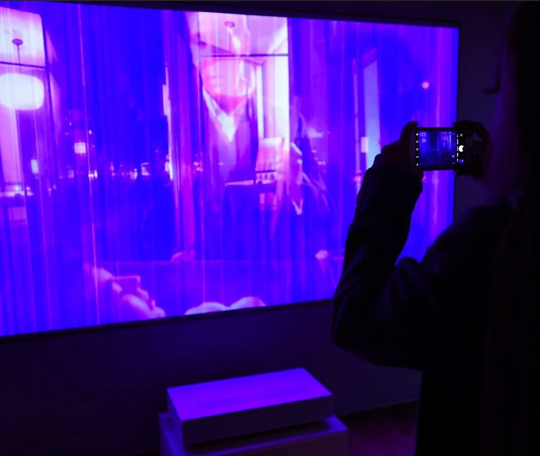 Person taking a photo of a brightly lit, purple-hued projection on a large screen in a dark room.