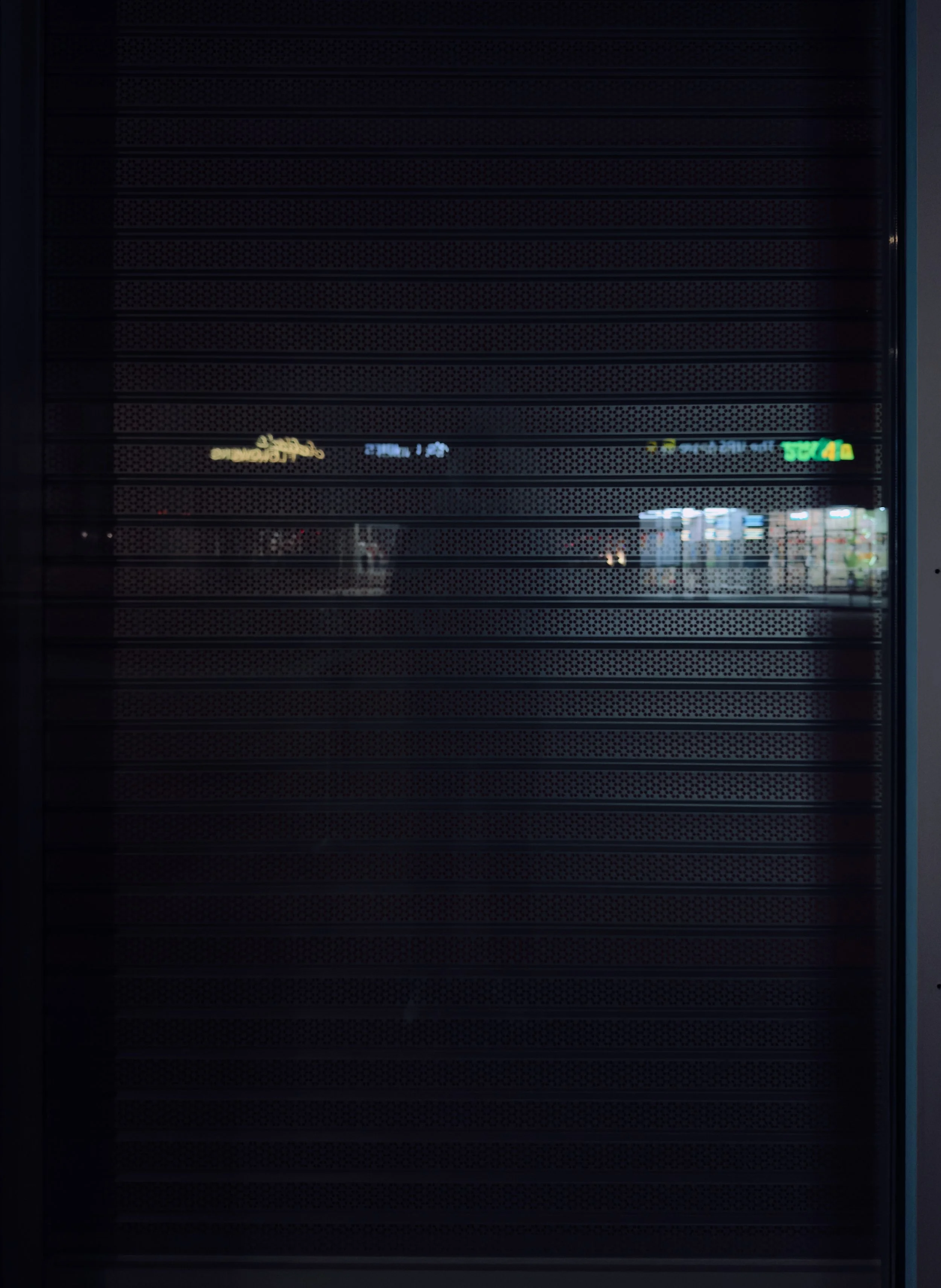 Nighttime view through a closed metal security shutter, with some illuminated signs and lights visible in the distance behind it.