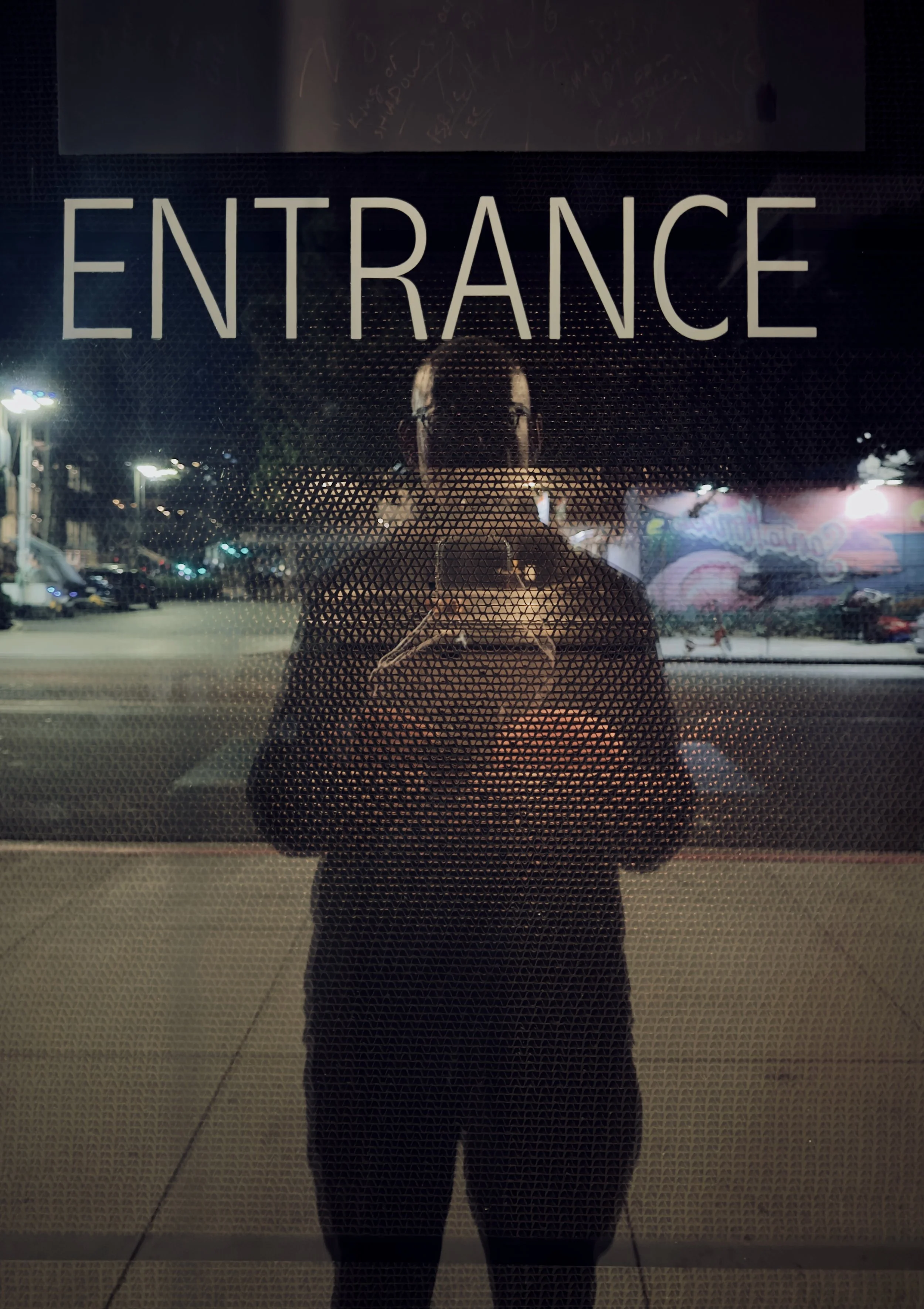 Nighttime exterior view of a glass door with a person taking a photo, and a sign that says 'ENTRANCE' in large letters.