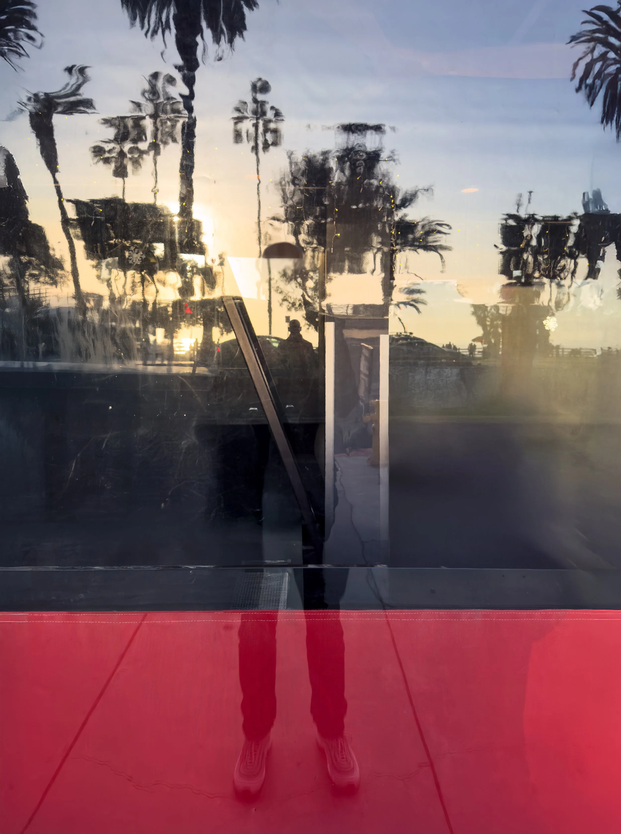 Reflection of a person wearing red pants and white shoes, standing outside near palm trees, with a sunset sky in the background, captured through a glass window.