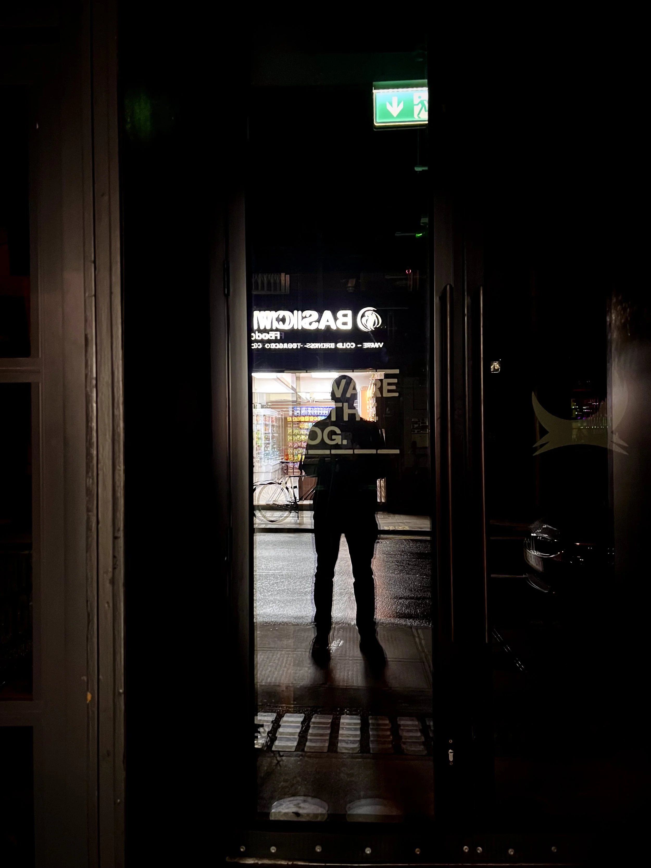 Reflection of a person taking a photo in a glass door with an illuminated store sign and exit sign above.