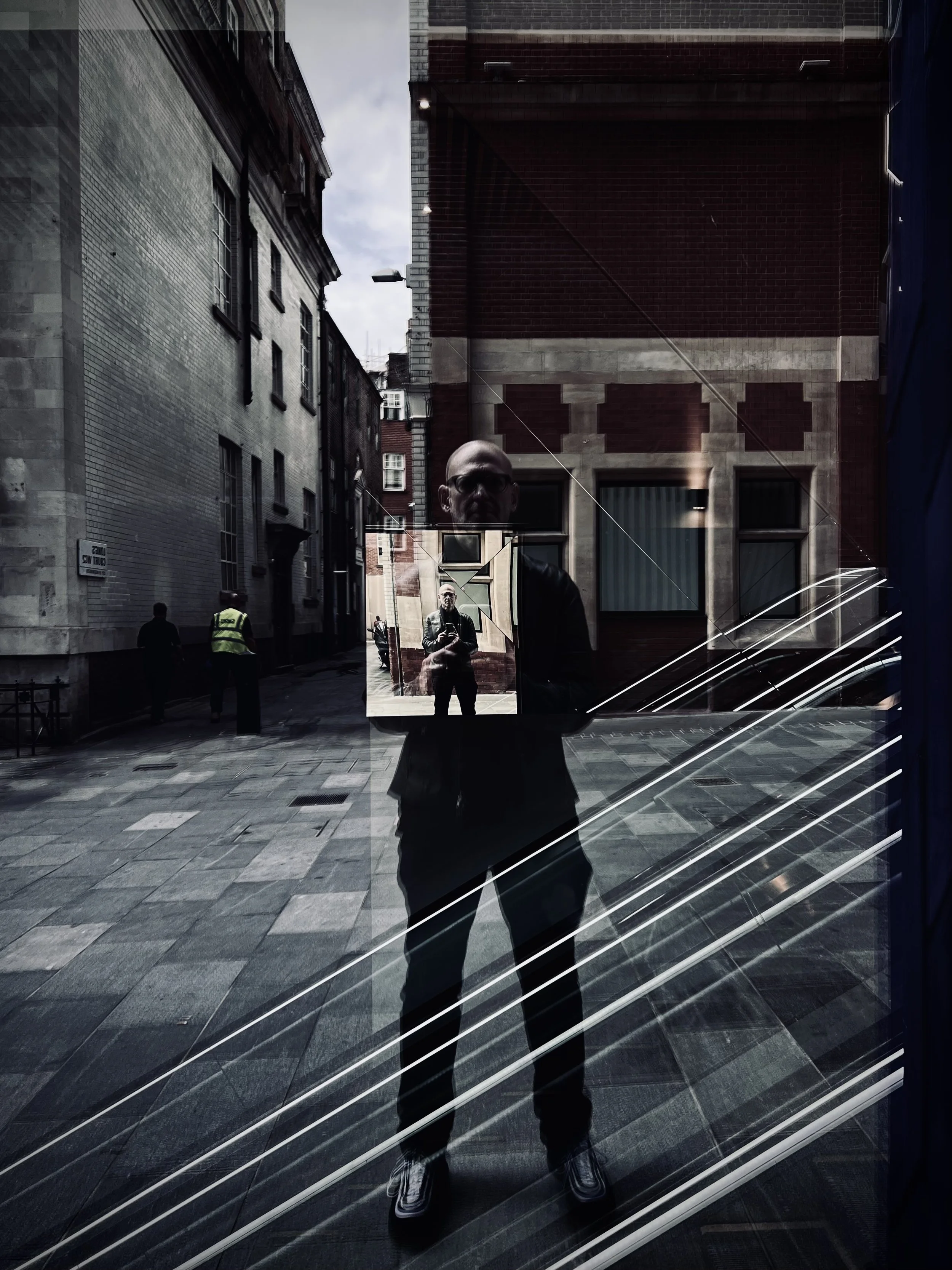 Reflection of a man taking a selfie in a window, with city buildings in the background and other pedestrians on the street.