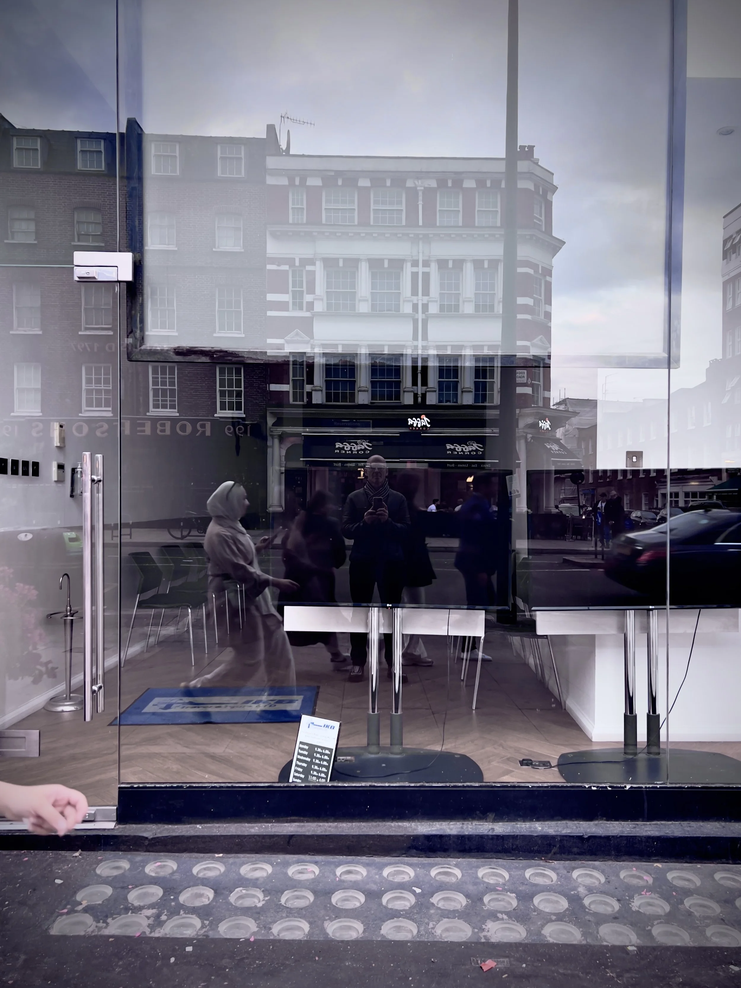 Reflection of people in a glass window of a storefront displaying a street scene with buildings and cars.