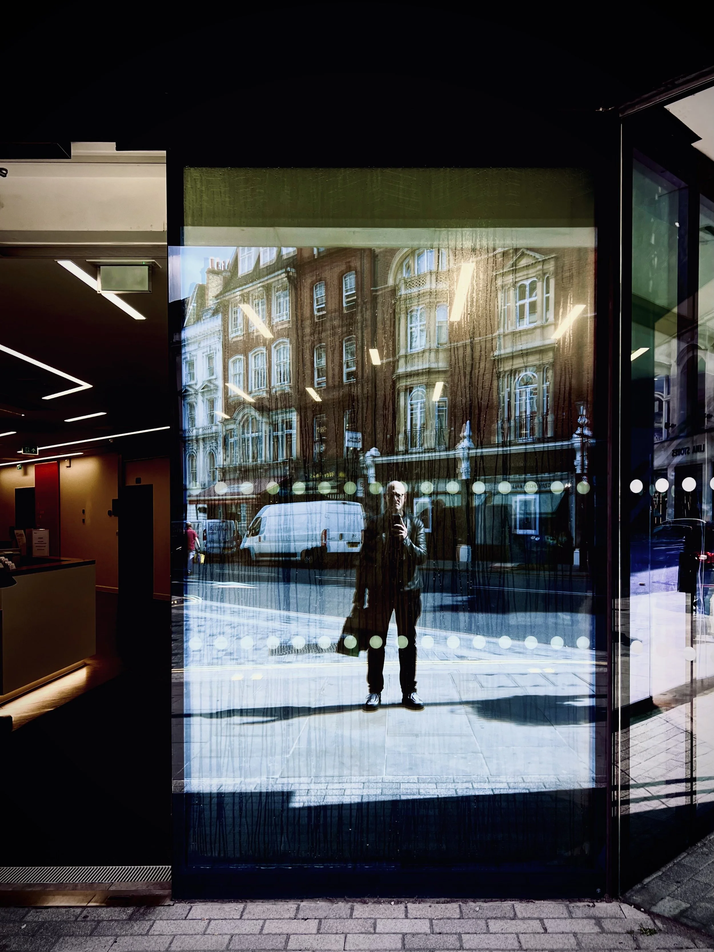 Reflection of a man taking a selfie in a glass door with city buildings and a van reflected in the glass.