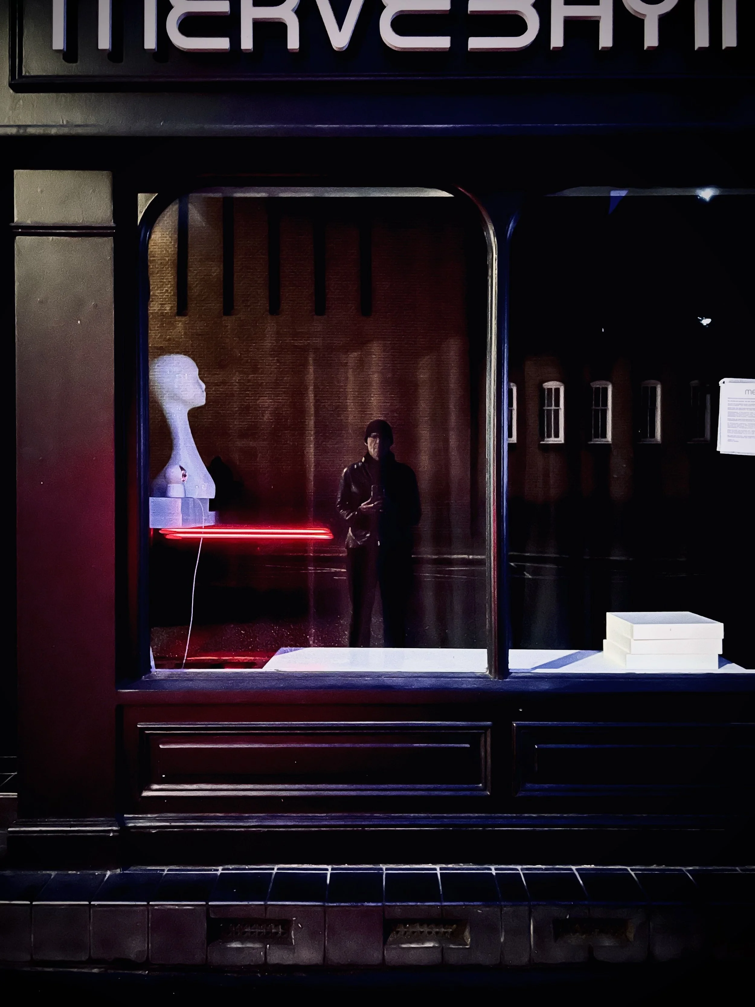 Storefront window display at night with a mannequin bust, a standing mannequin, red neon light, and white boxes.