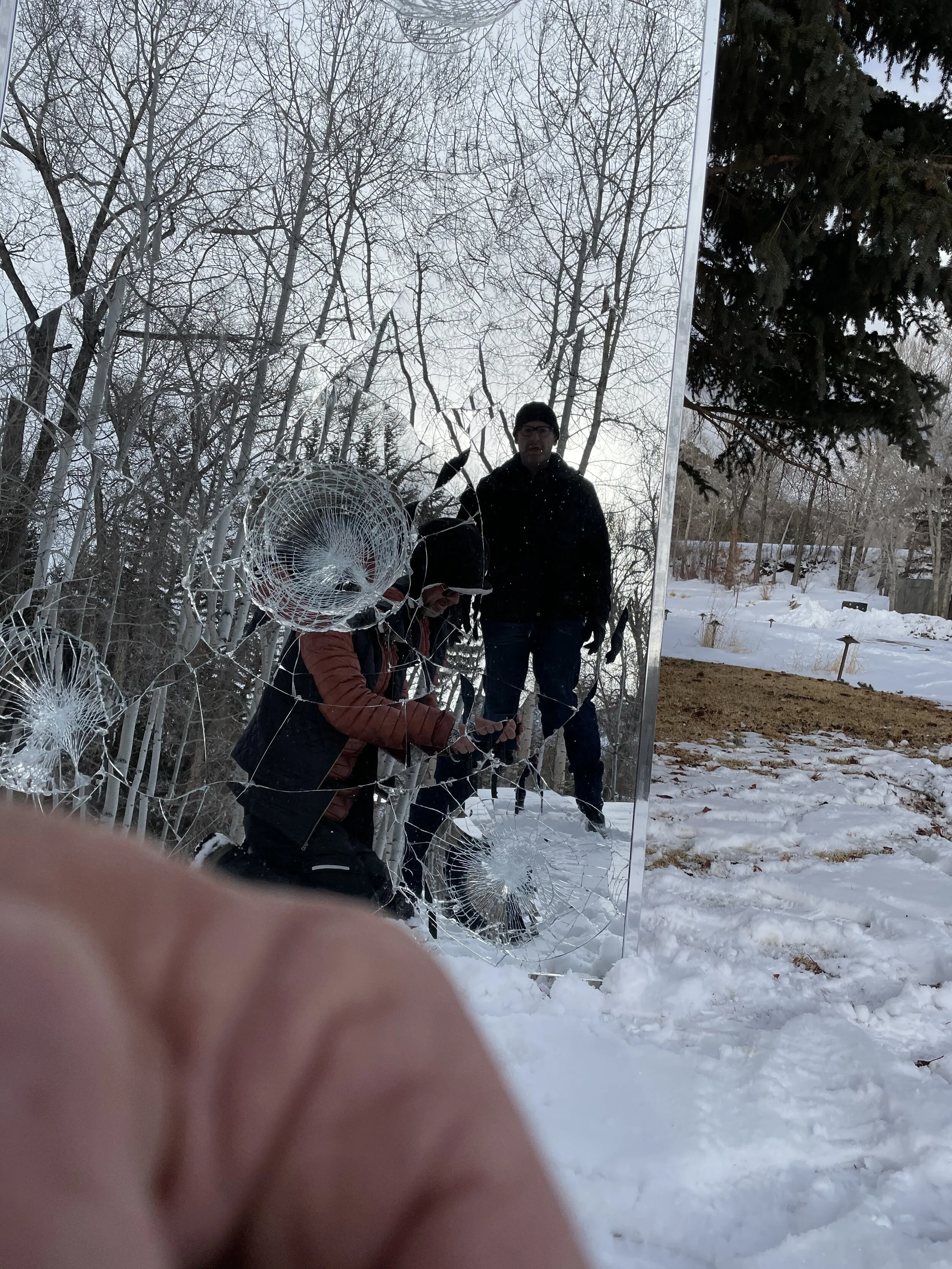 People taking photos of a cracked mirror in a snowy landscape with bare trees and a fir tree.