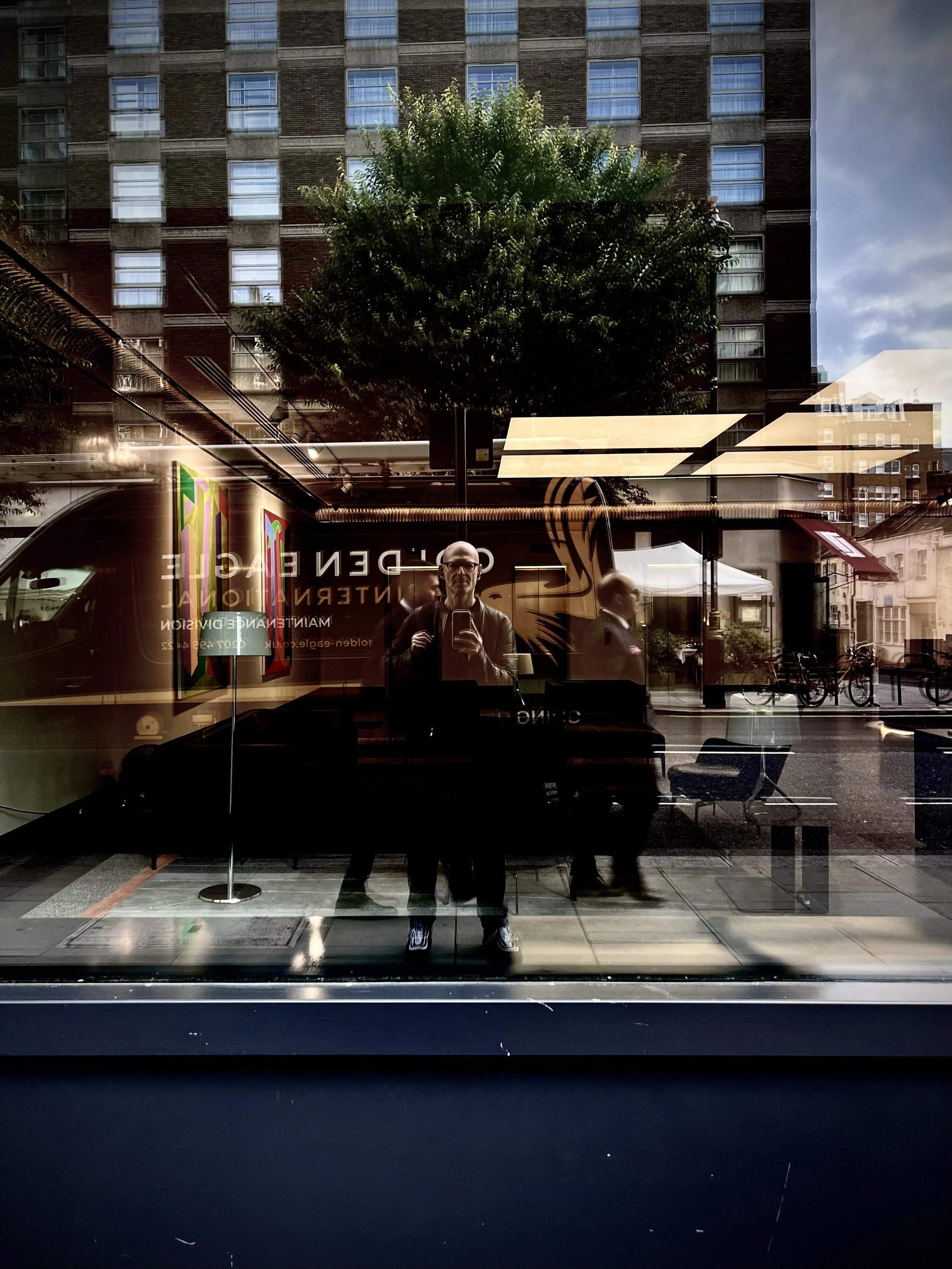 Reflection of a man taking a photo in a glass window with city buildings, a tree, and outdoor seating visible in the background.