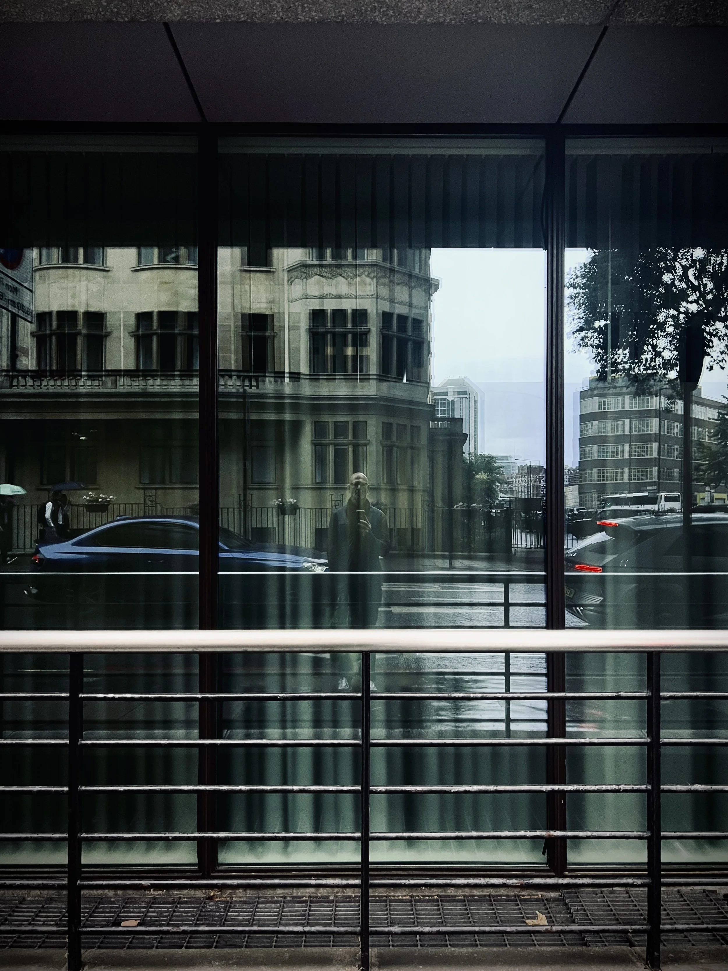 City street scene reflected in a glass window with railings in the foreground, an ornate building across the street, and cars passing by.