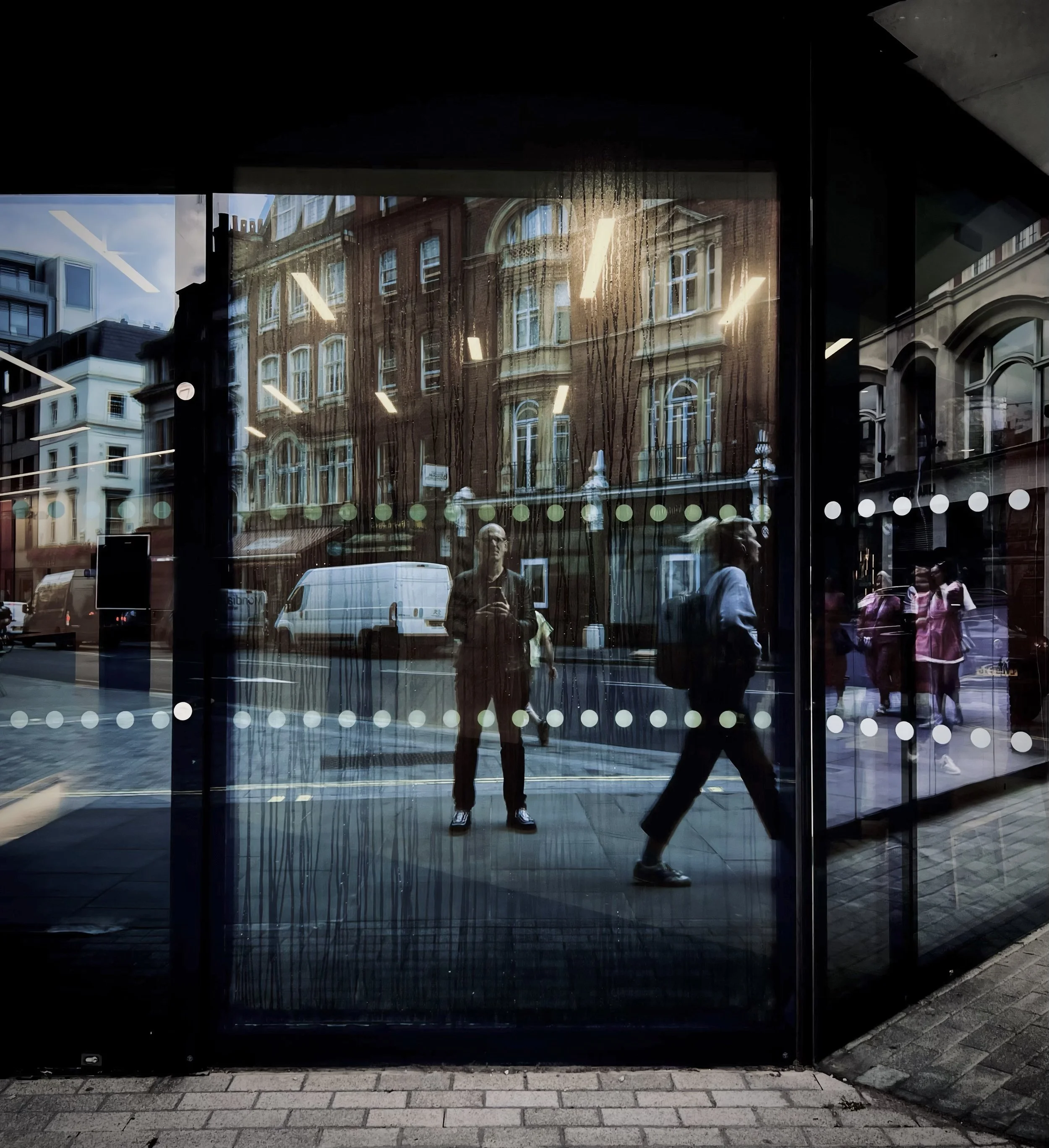 Reflection of city street scene with buildings, cars, and pedestrians seen through glass window with white dots, including a person taking a photo.