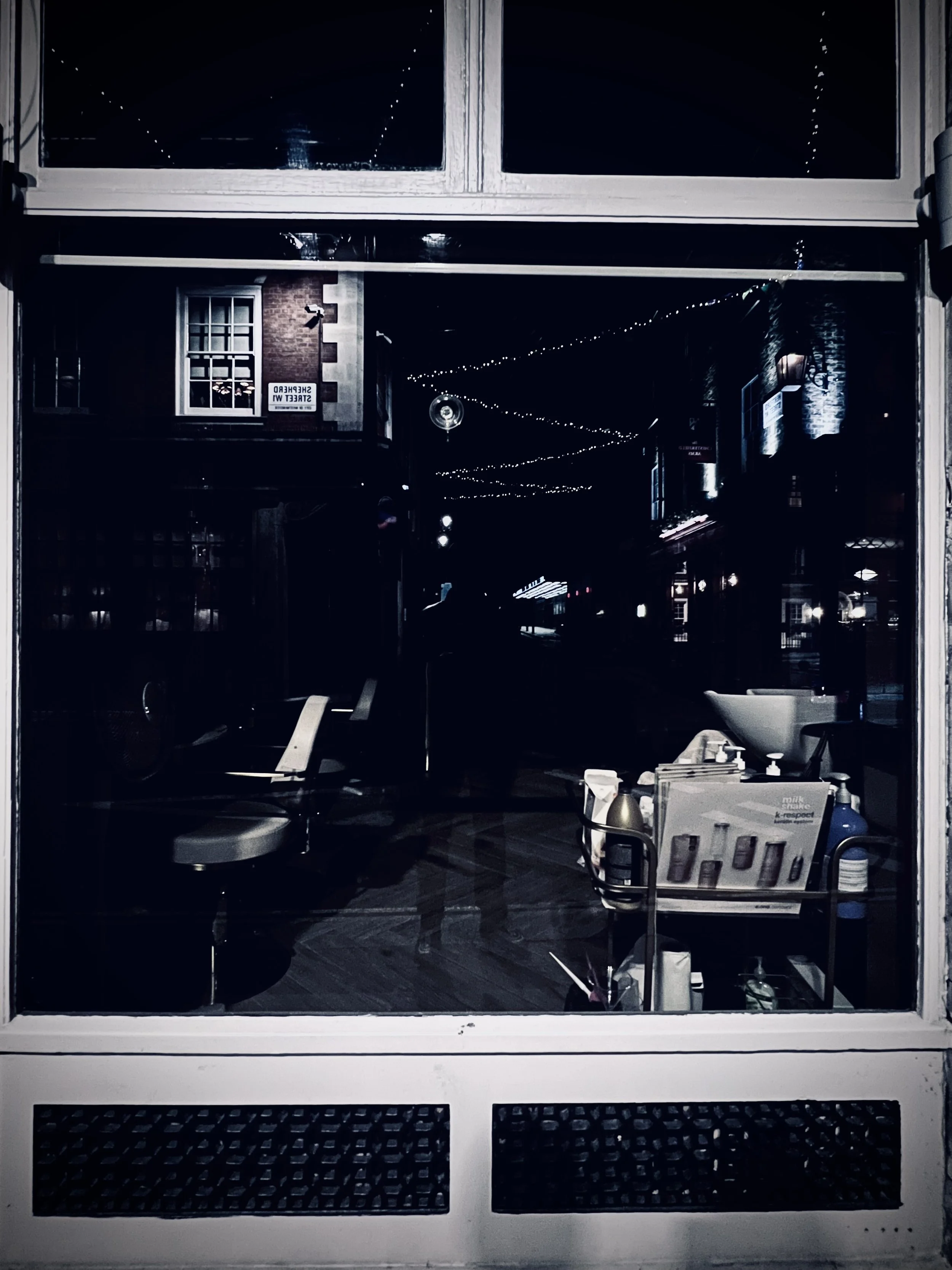 View through a window into a dimly lit salon at night, showing salon chairs, a cart with hair care products, and a bowl sink; reflections of the street outside are visible.