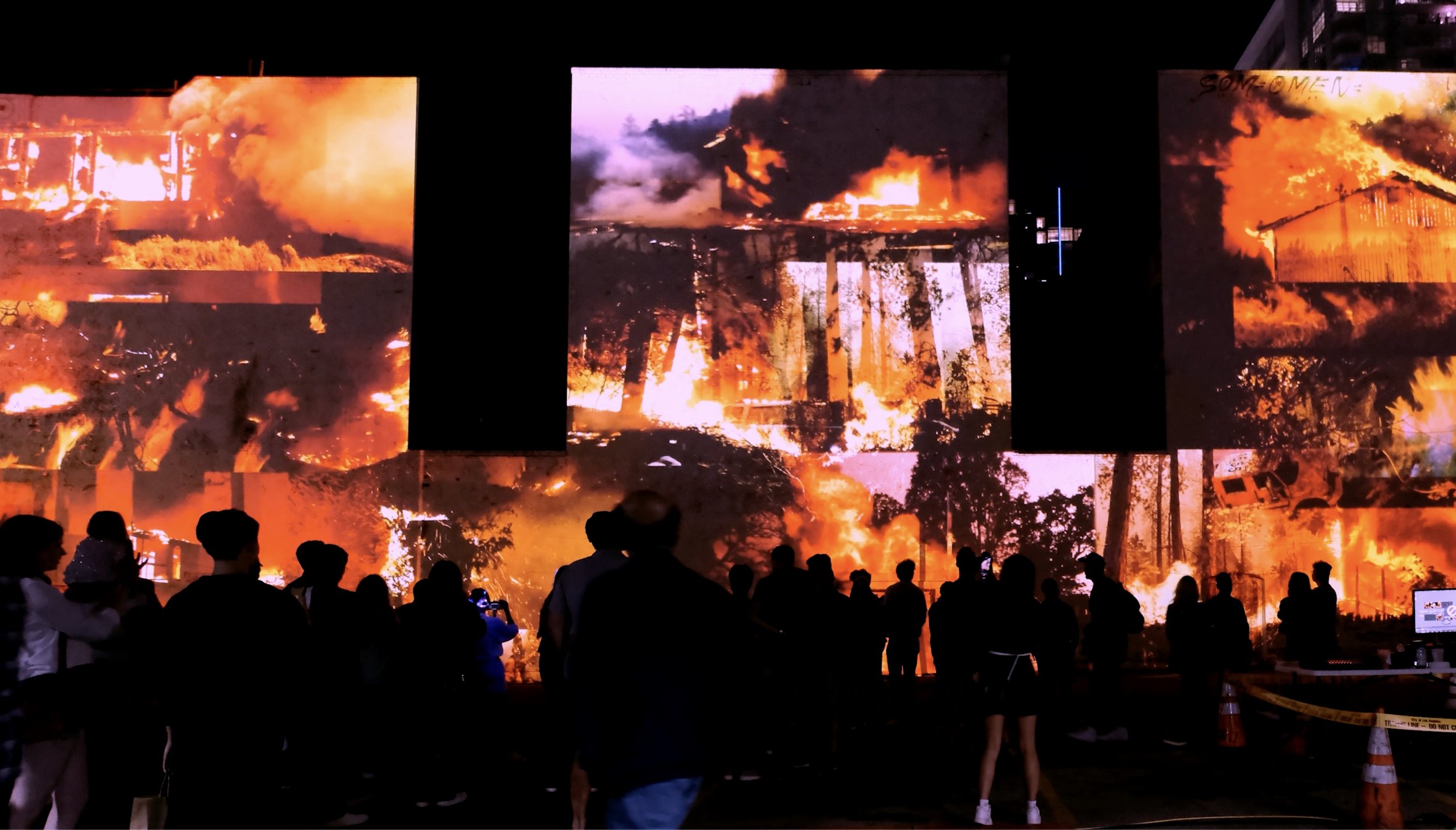 Crowd watching large outdoor screens displaying fire scenes at night.