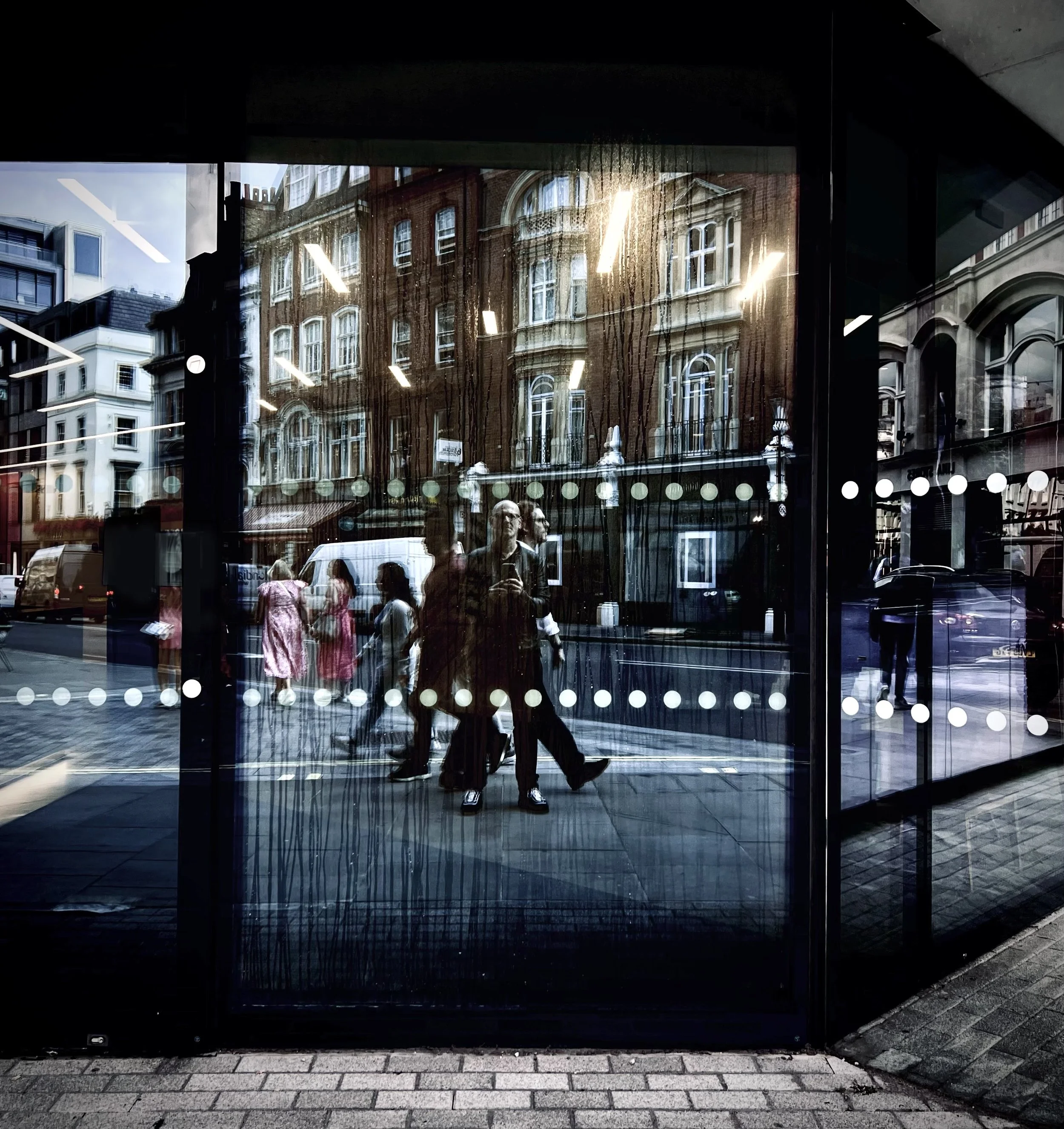 Reflection of people walking on city street seen through glass window with white dotted line design, showing buildings and pedestrians outside.