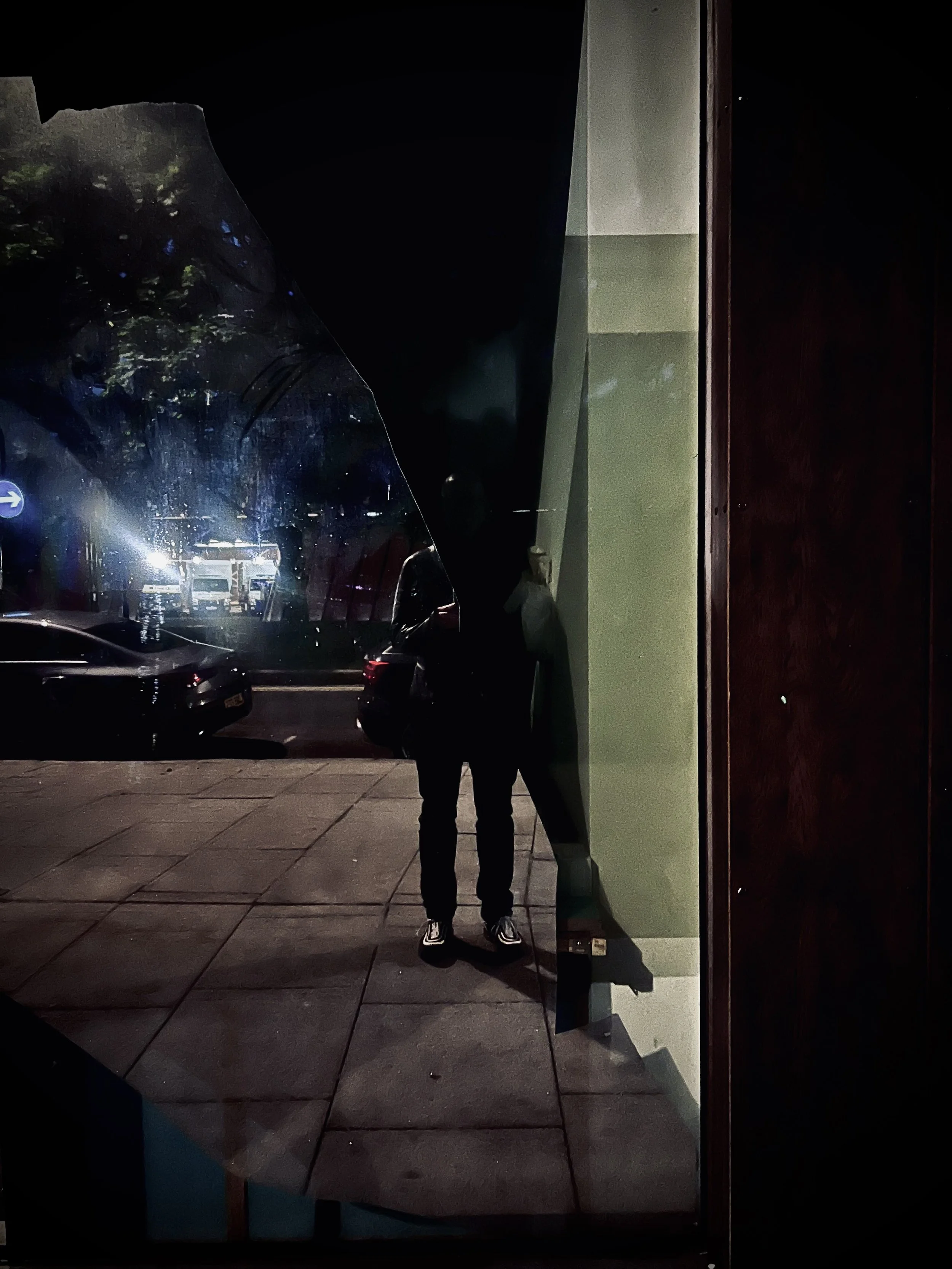 Nighttime street scene reflected in a window, showing parked cars and a person taking a photo.