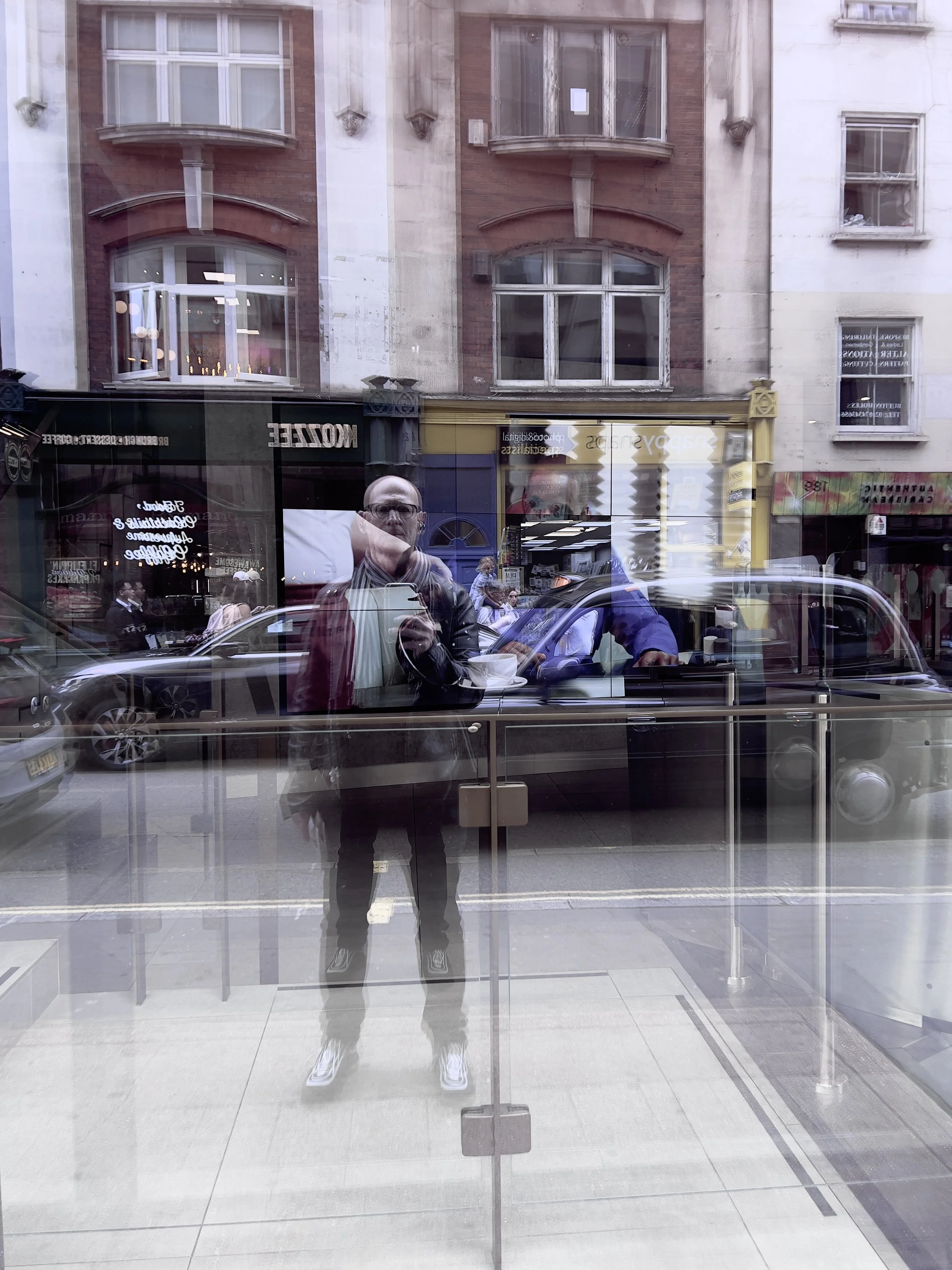 Reflection of a man taking a photo in a glass window with city street and buildings in the background.