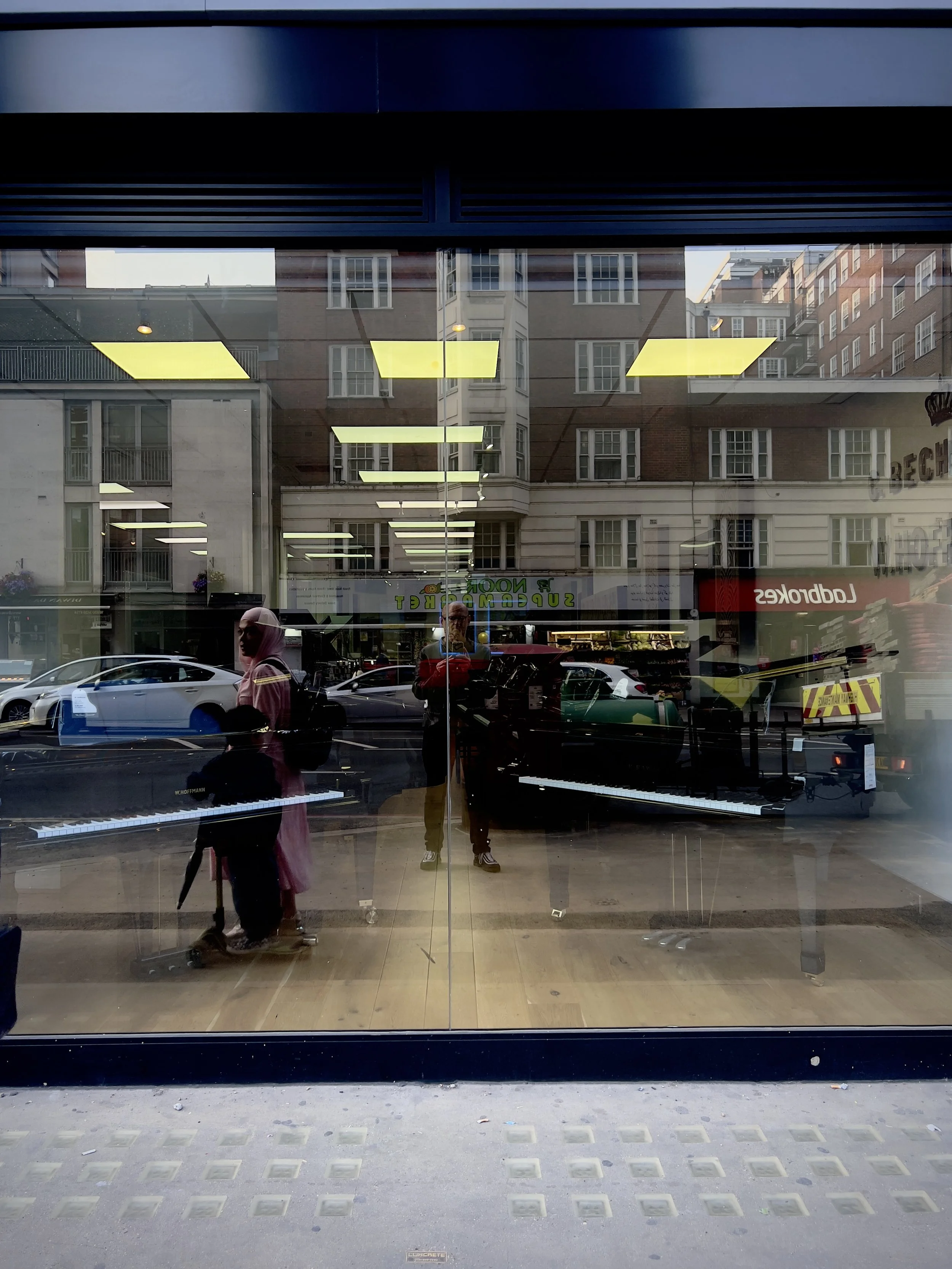 Reflection of a person taking a photo inside a storefront window, with a woman playing a keyboard and various cars and buildings outside