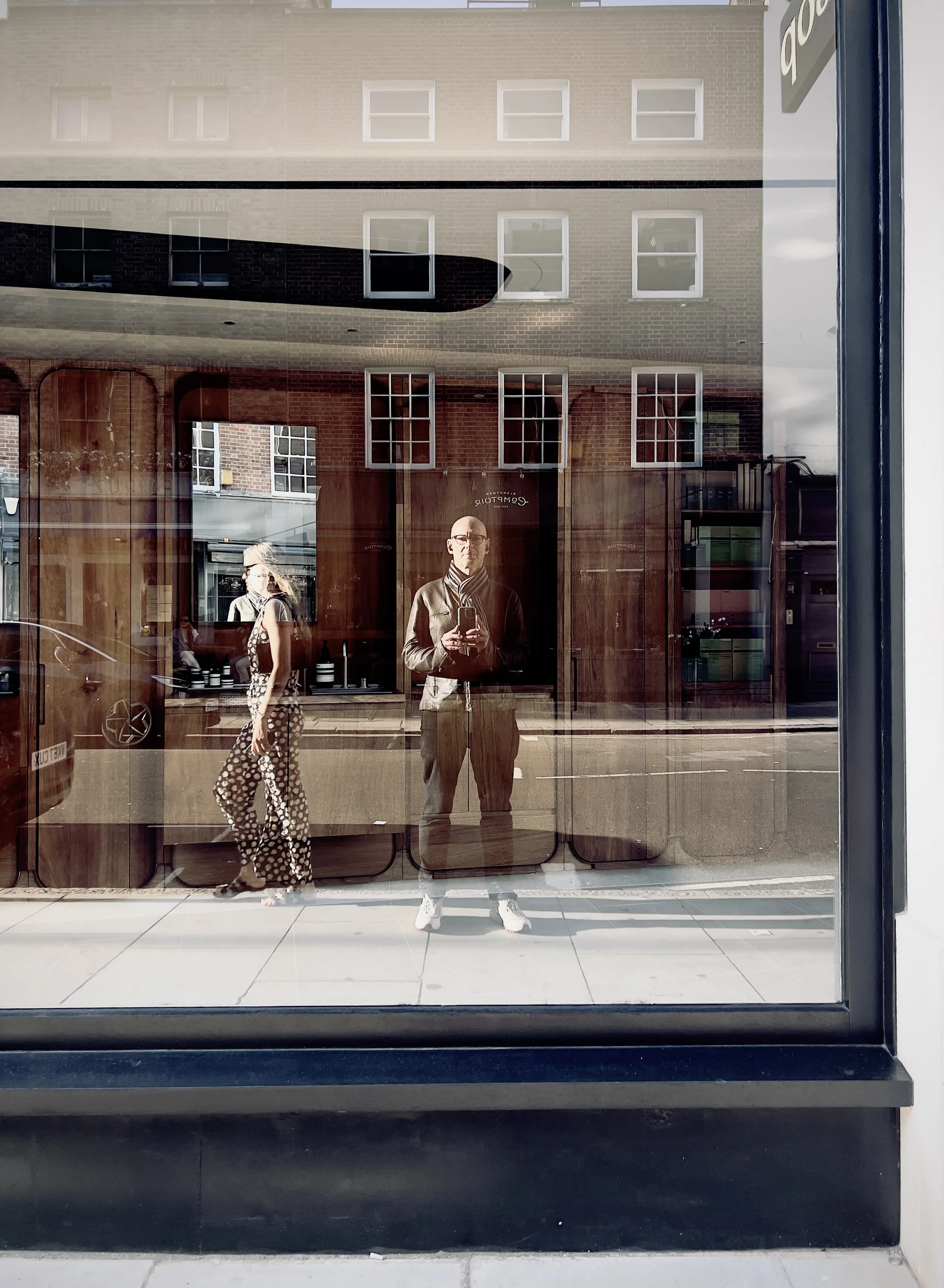 Reflection of two pedestrians and a man taking a selfie seen in store window with glass storefront