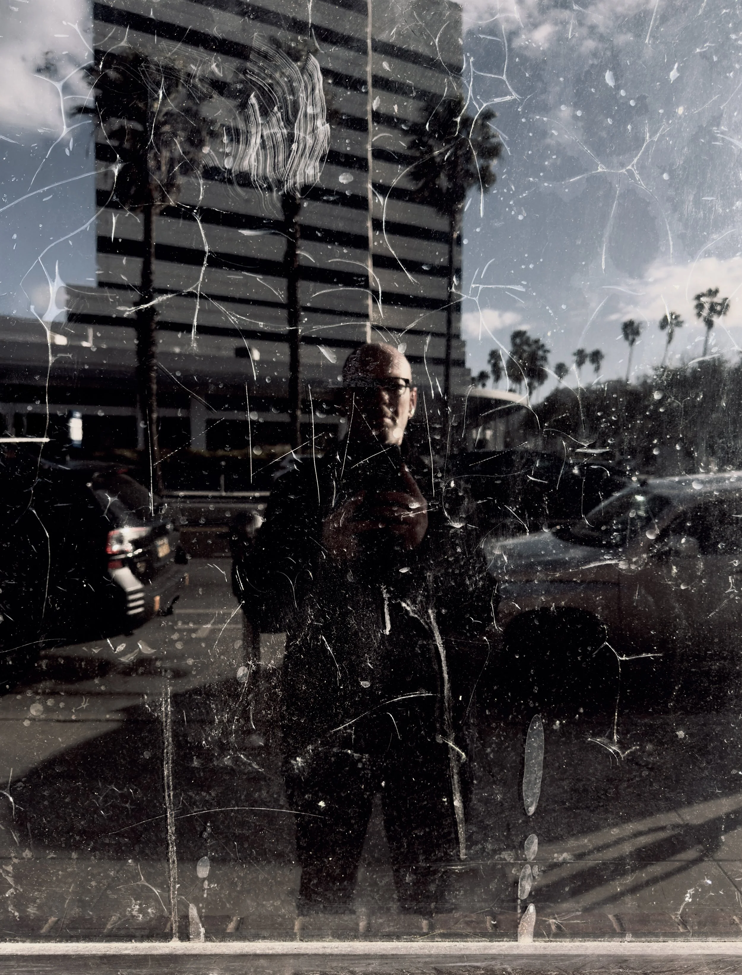 Person taking a photo of their reflection on a dirty glass window with scratches, in an urban parking lot with buildings, cars, palm trees, and a partly cloudy sky in the background.