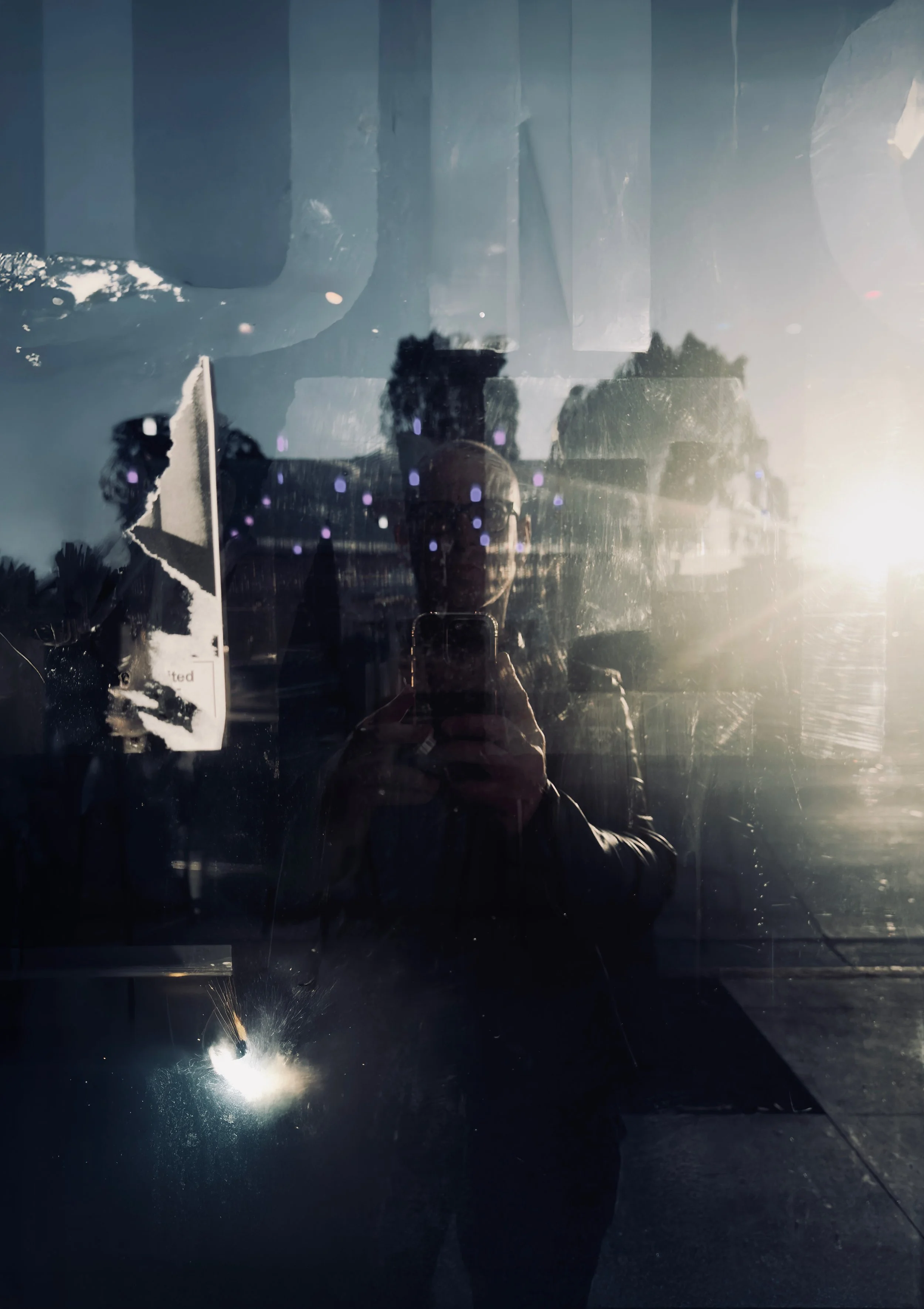Reflection of a person taking a photo in a smudged glass window with sunlight in the background.