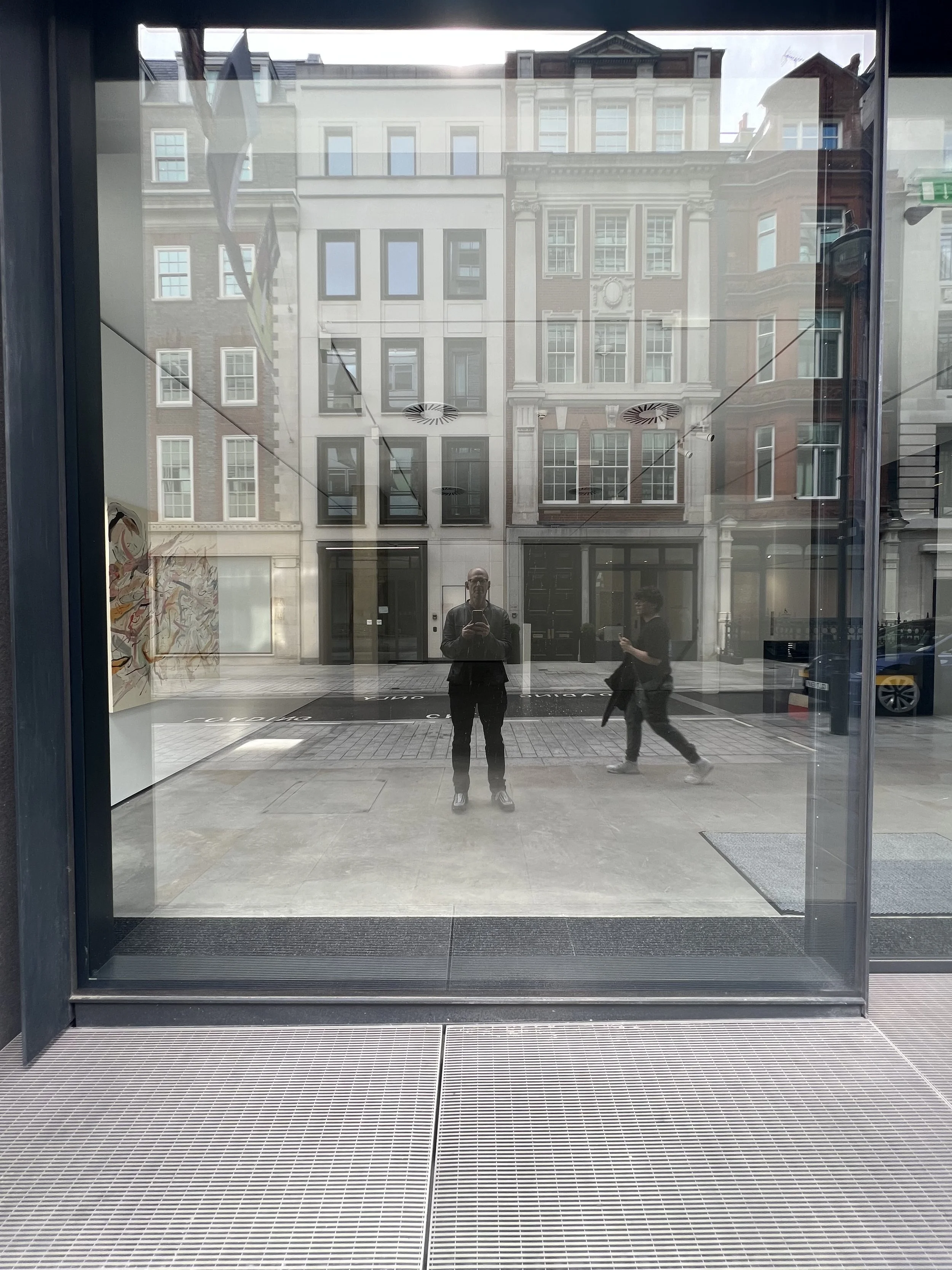 Reflection of a person taking a photo in a large glass window, with pedestrians and buildings visible across the street.