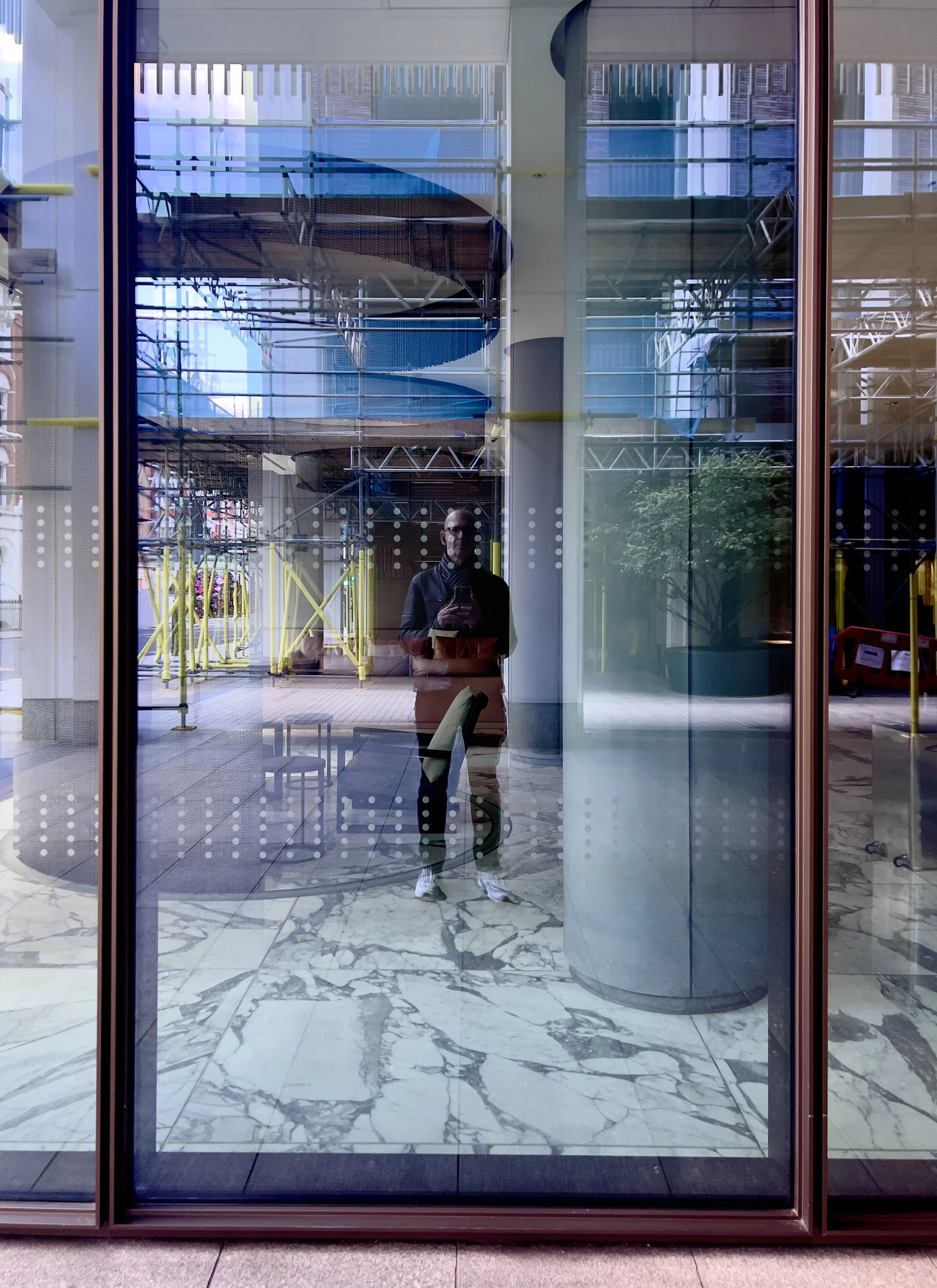 Reflection of a person taking a photo in a glass window, with a modern indoor/outdoor space, colorful slide, yellow structural elements, chairs, and marble floor visible.