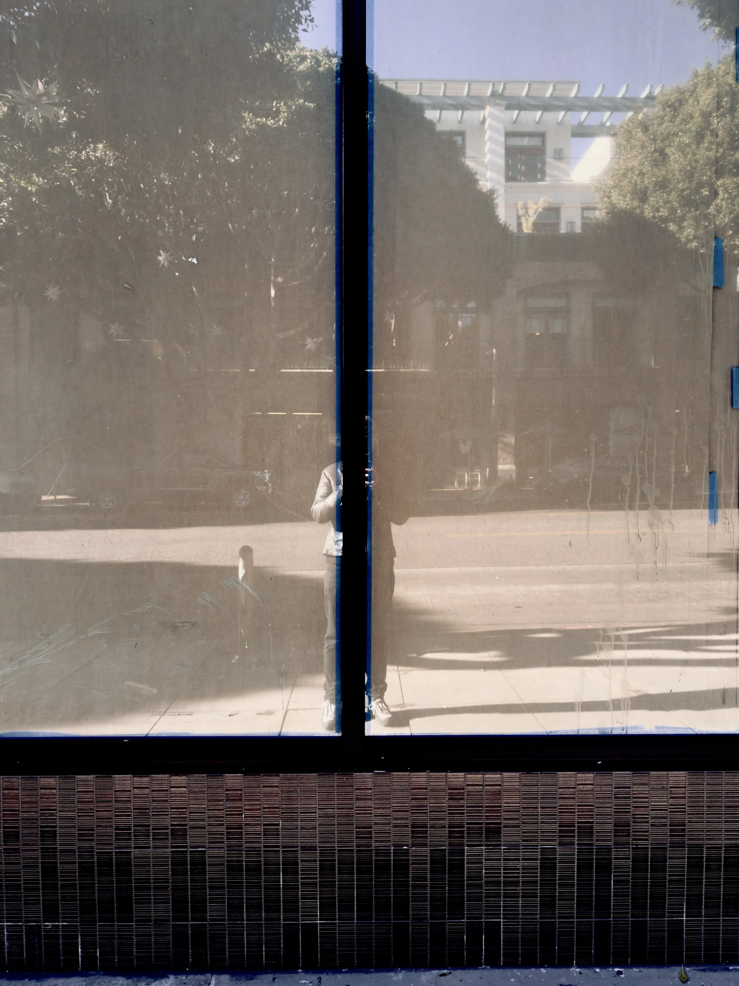 Reflection of a person with white shoes and a white jacket taking a photo in front of glass doors, with trees and a building visible outside.