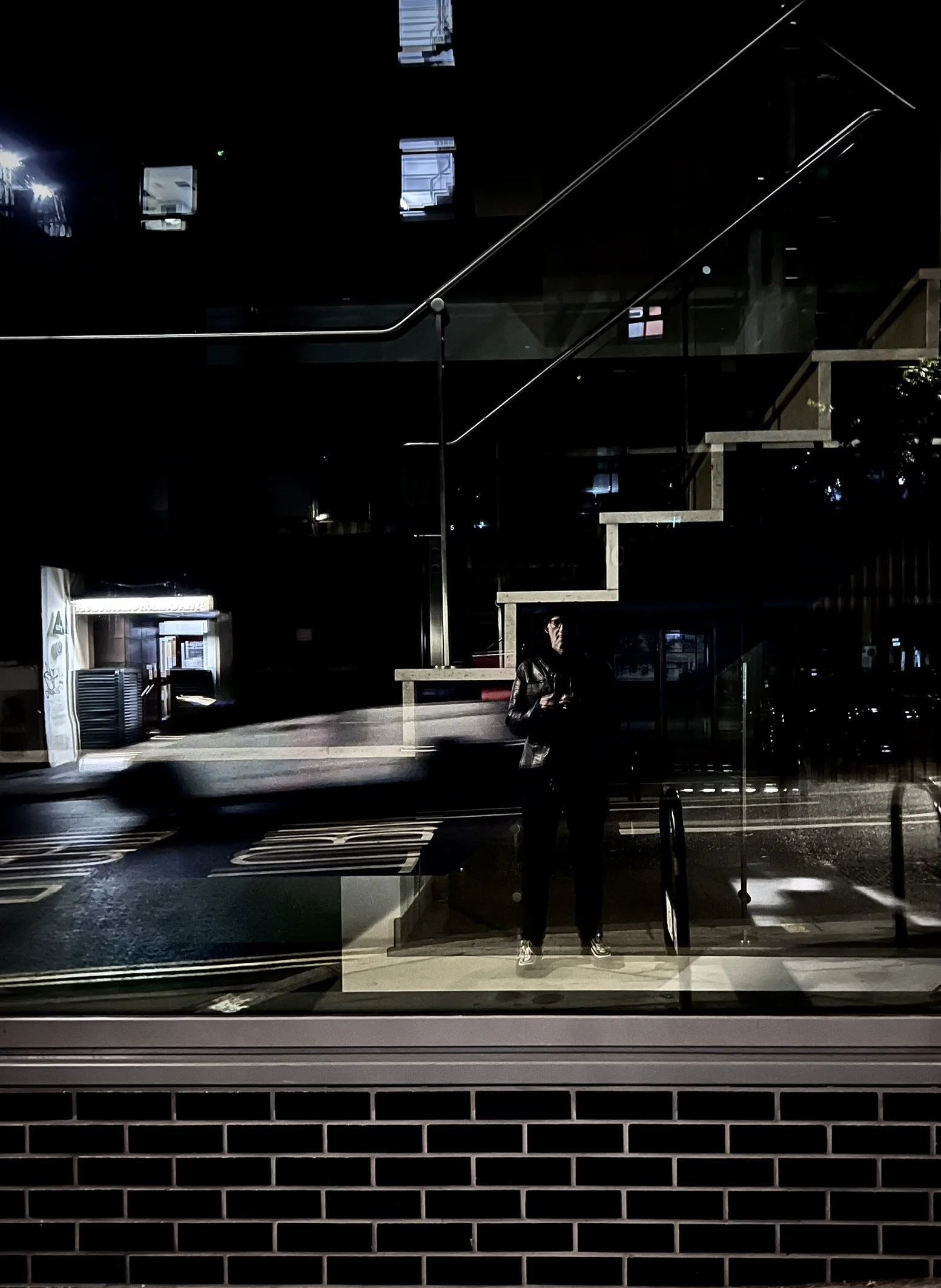 A person taking a photo of their reflection in a dark glass window at night. The scene includes an exterior brick wall at the bottom and a staircase and building reflections in the background.