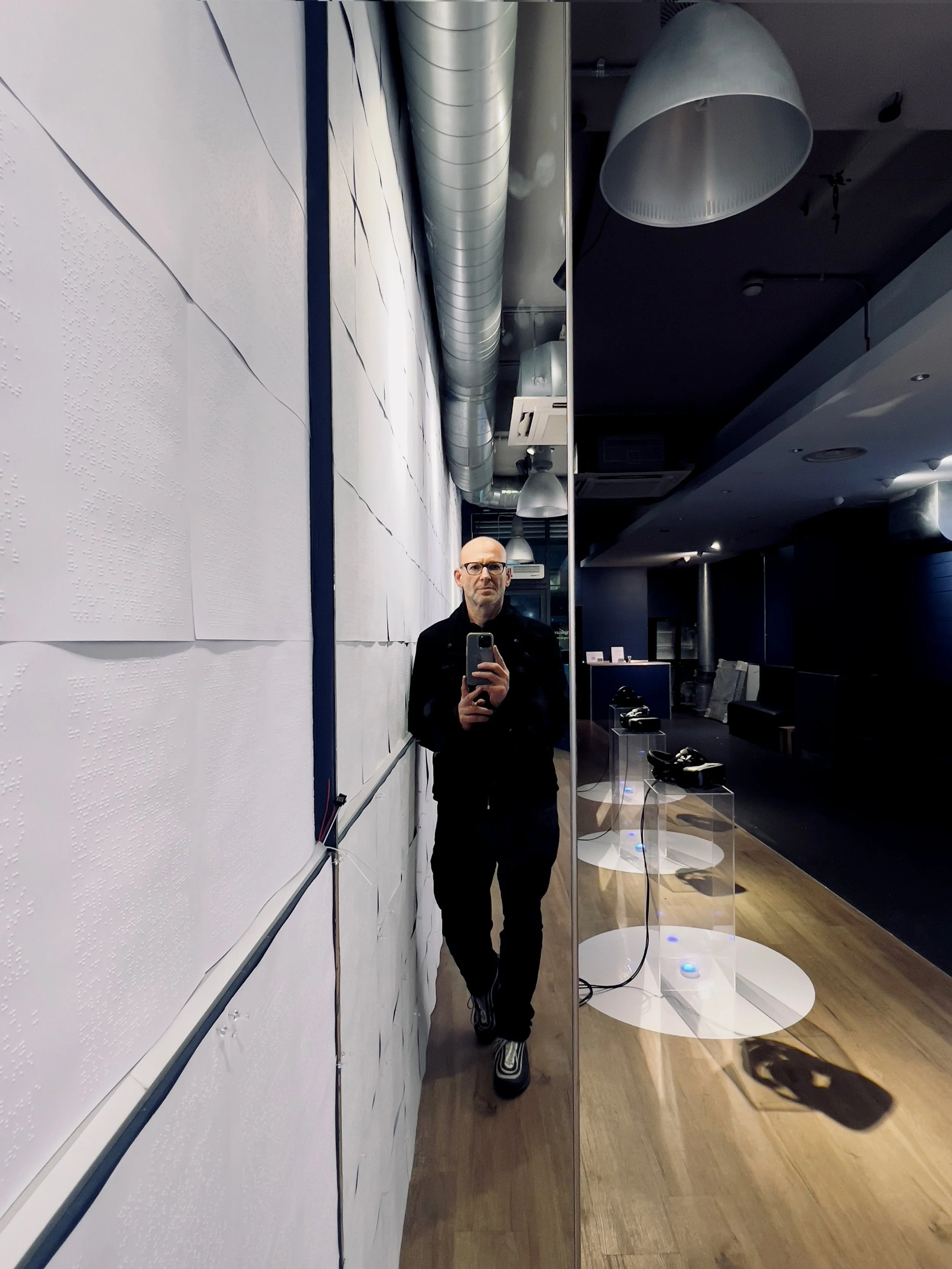 Person taking a mirror selfie in a modern room with white walls and a wooden floor. Several telephones are displayed on transparent stands.