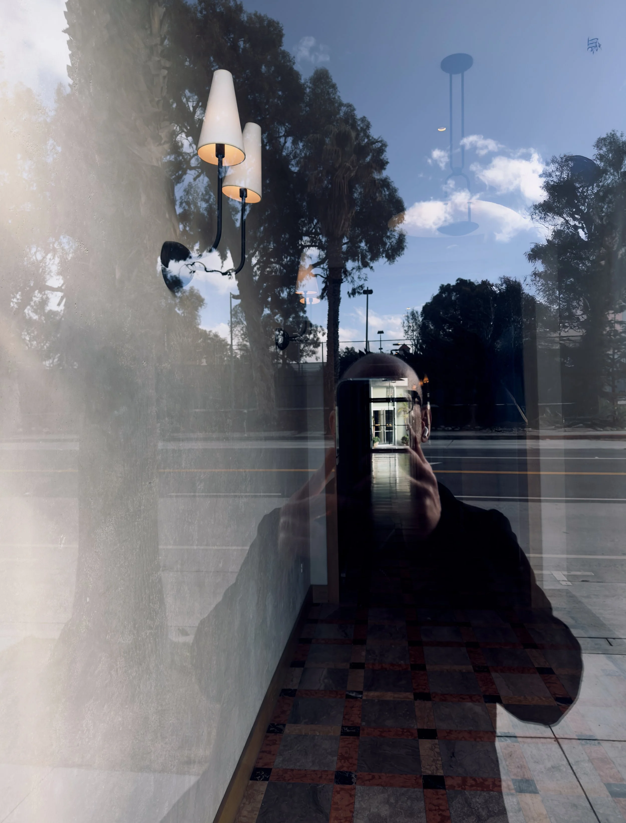 A reflection on a glass window showing a person taking a photo indoors, with a view of the sky, trees, and a chandelier outside.