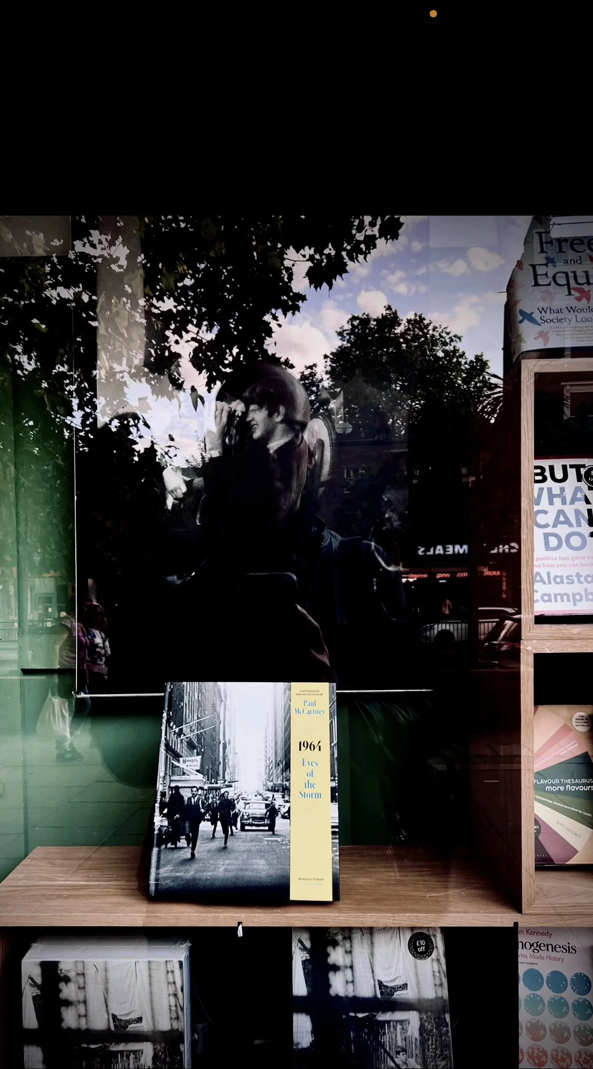 A book titled '1964 Eyes of the Storm' by Paul McCartney displayed on a wooden shelf, with a reflection of people and trees outside in the glass window behind it.