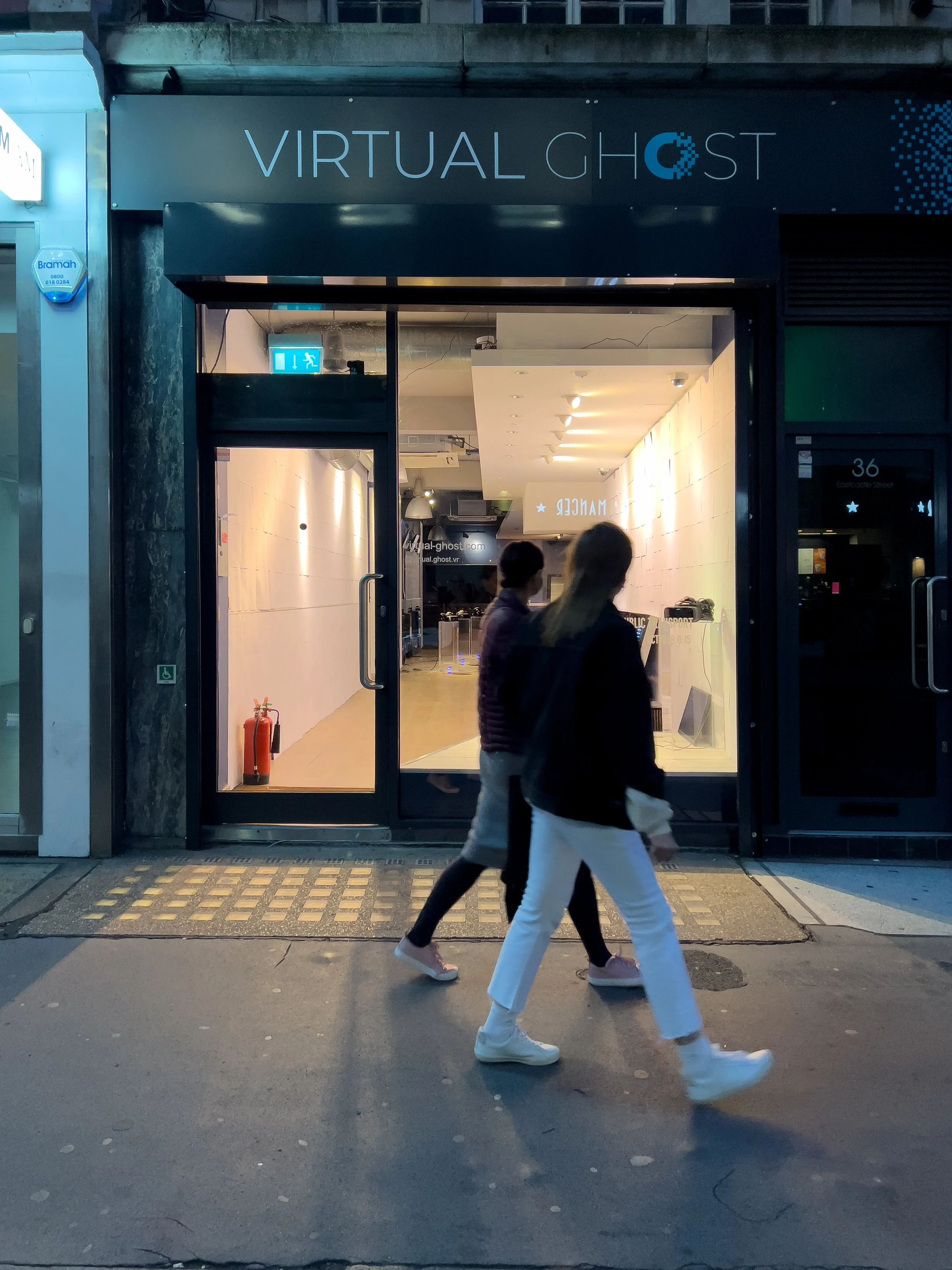 Facade of a storefront labeled "Virtual Ghost," with two people walking past on the sidewalk.