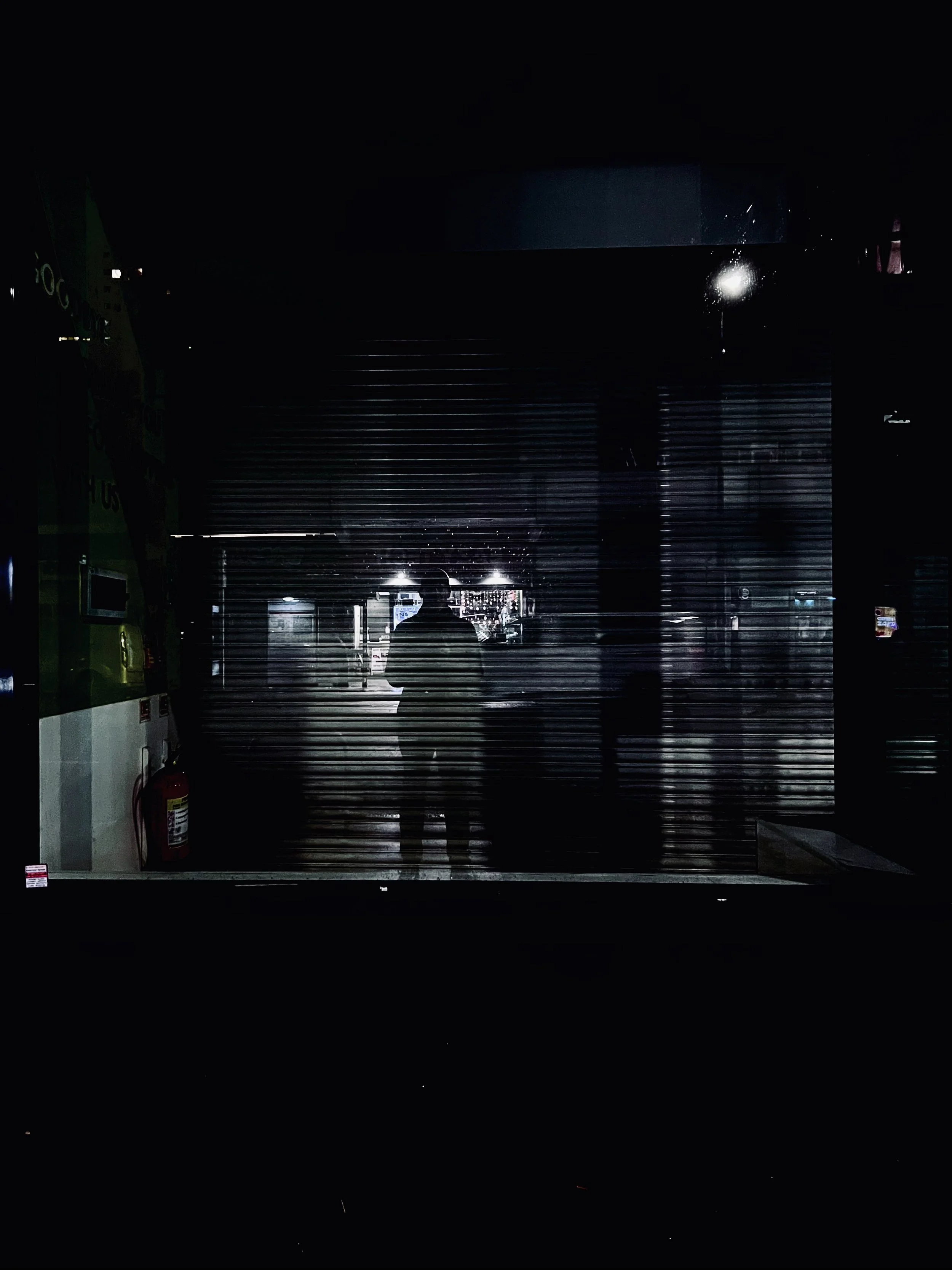 A silhouette of a person standing in front of a closed metal shutter at night, with minimal light reflecting off the shutter and surroundings.