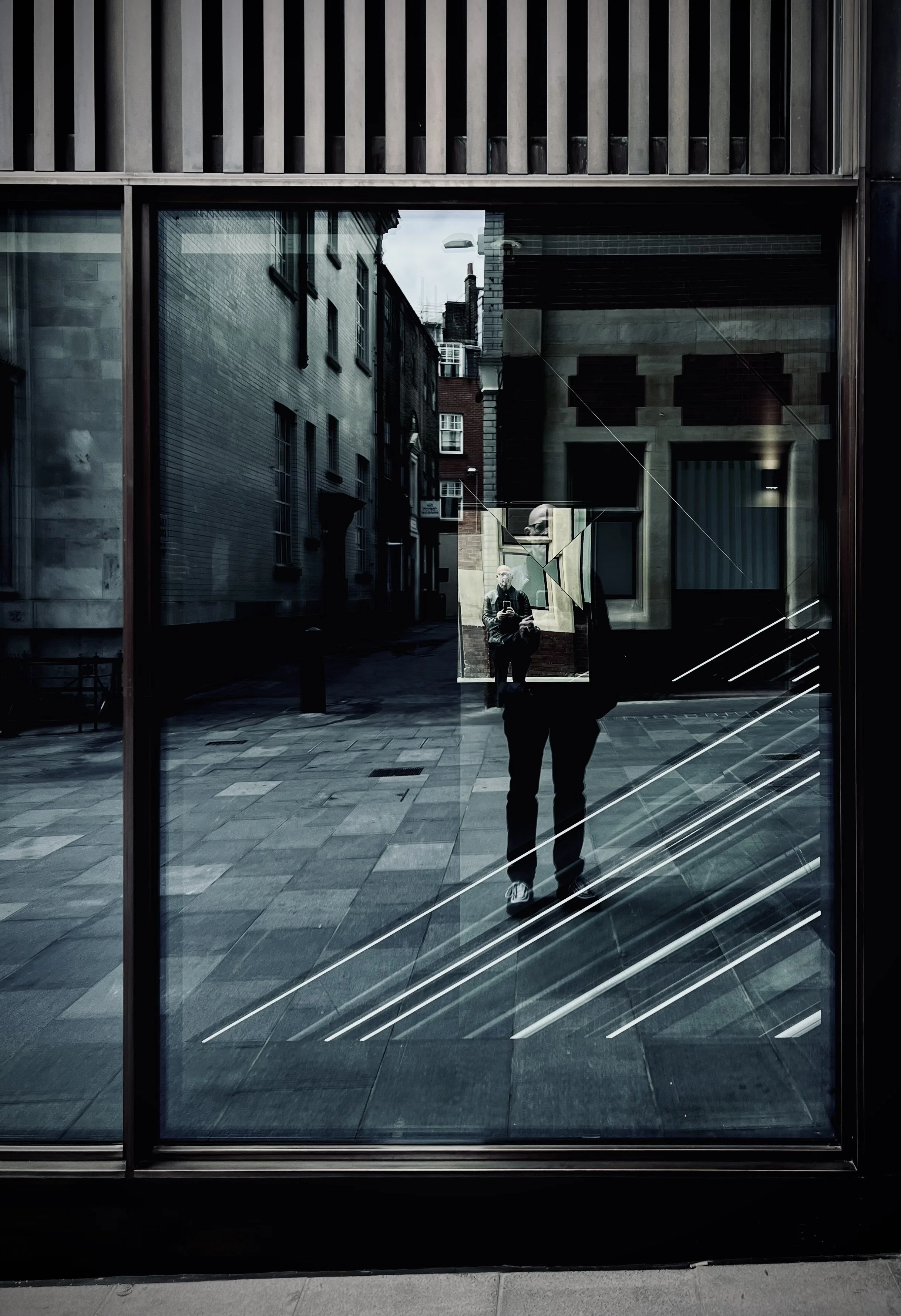 Reflection of a person taking a photo, seen through a large glass window, with an urban street scene in the background.