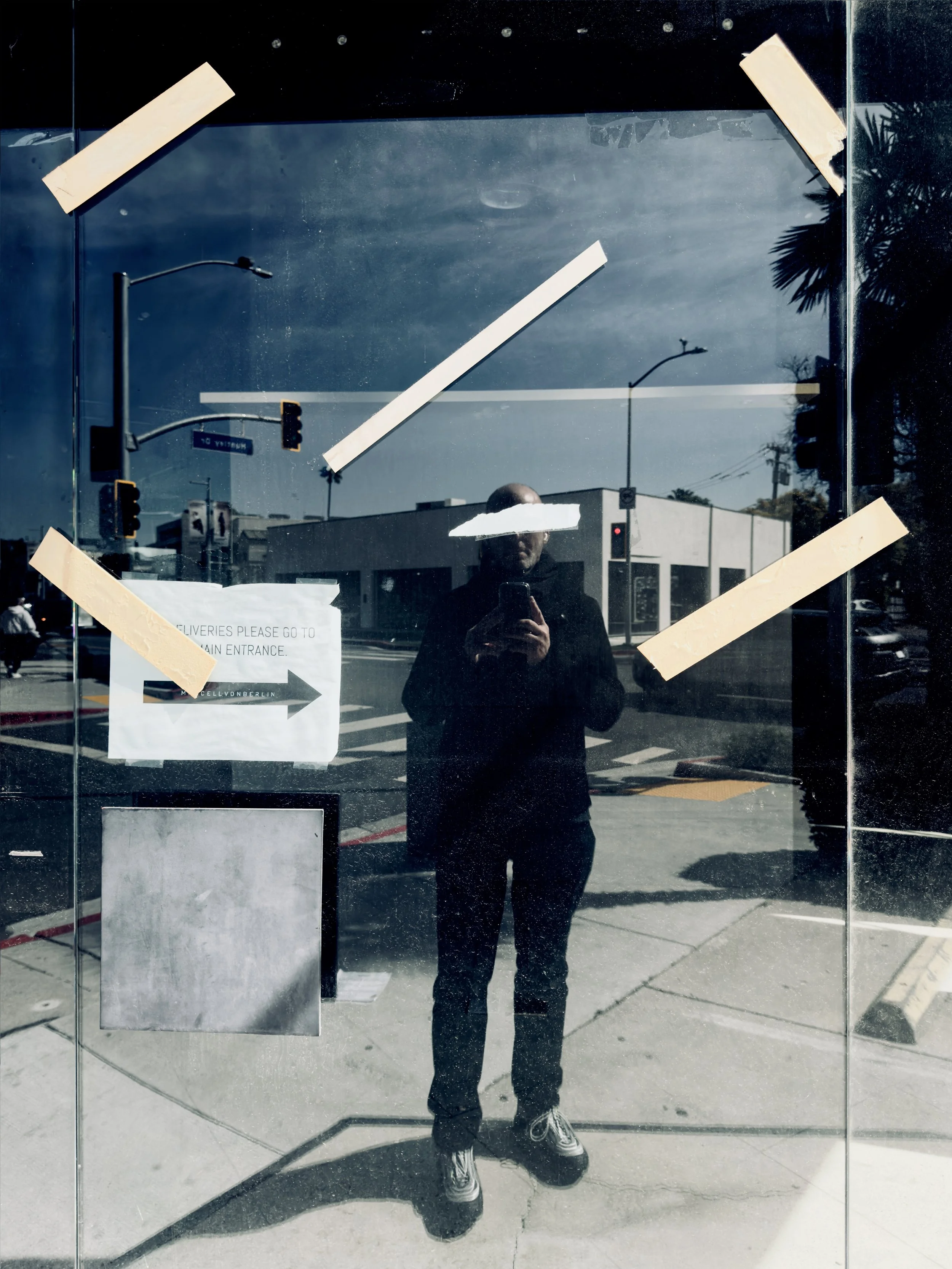 Reflection of a person taking a photo on a glass door or window with taped notes and a weathered sign, in an urban street scene with buildings, traffic lights, and pedestrians.