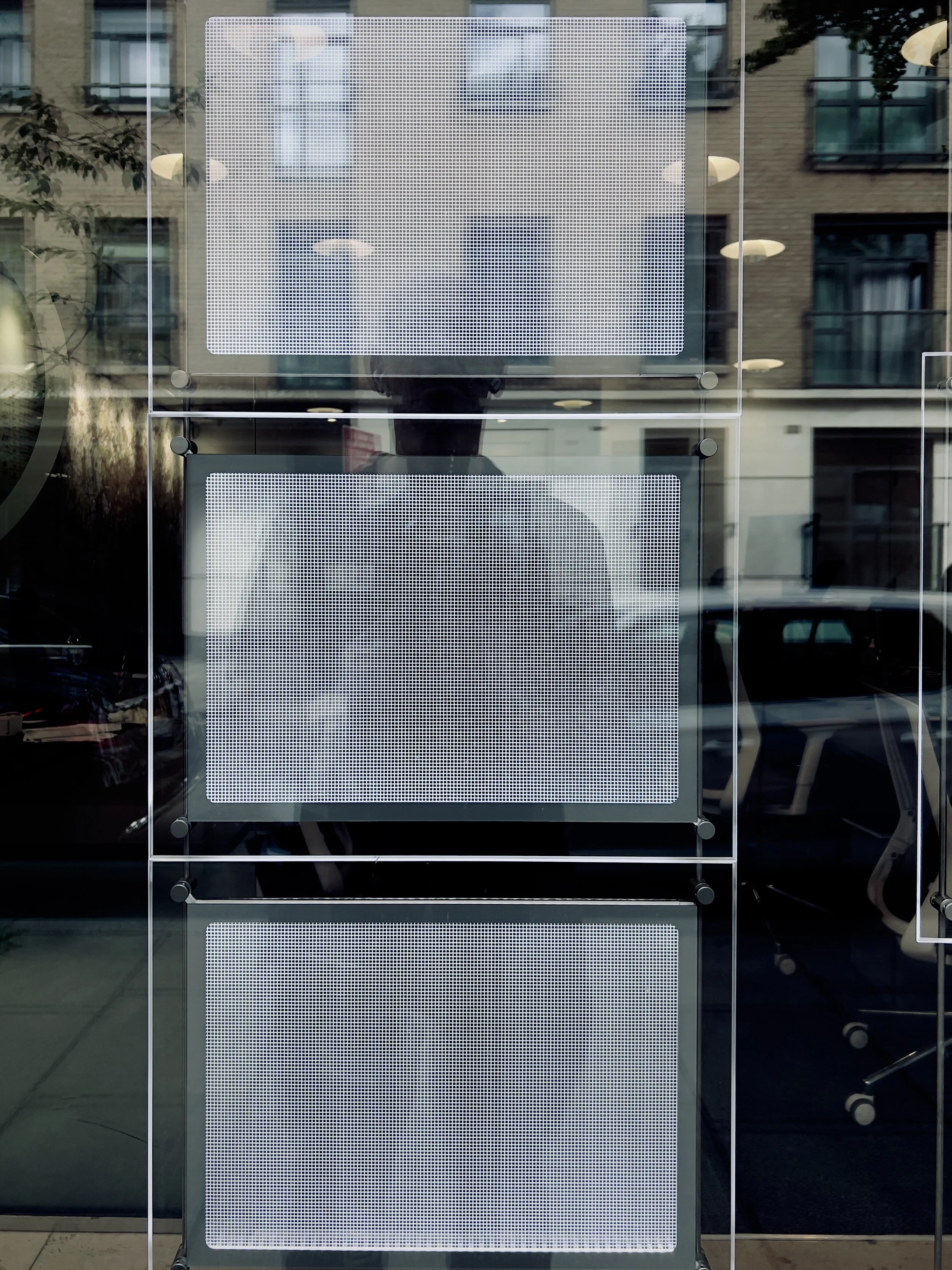 Three empty electronic display screens mounted on a glass window, with reflections of a street scene including buildings, cars, and indoor lights.