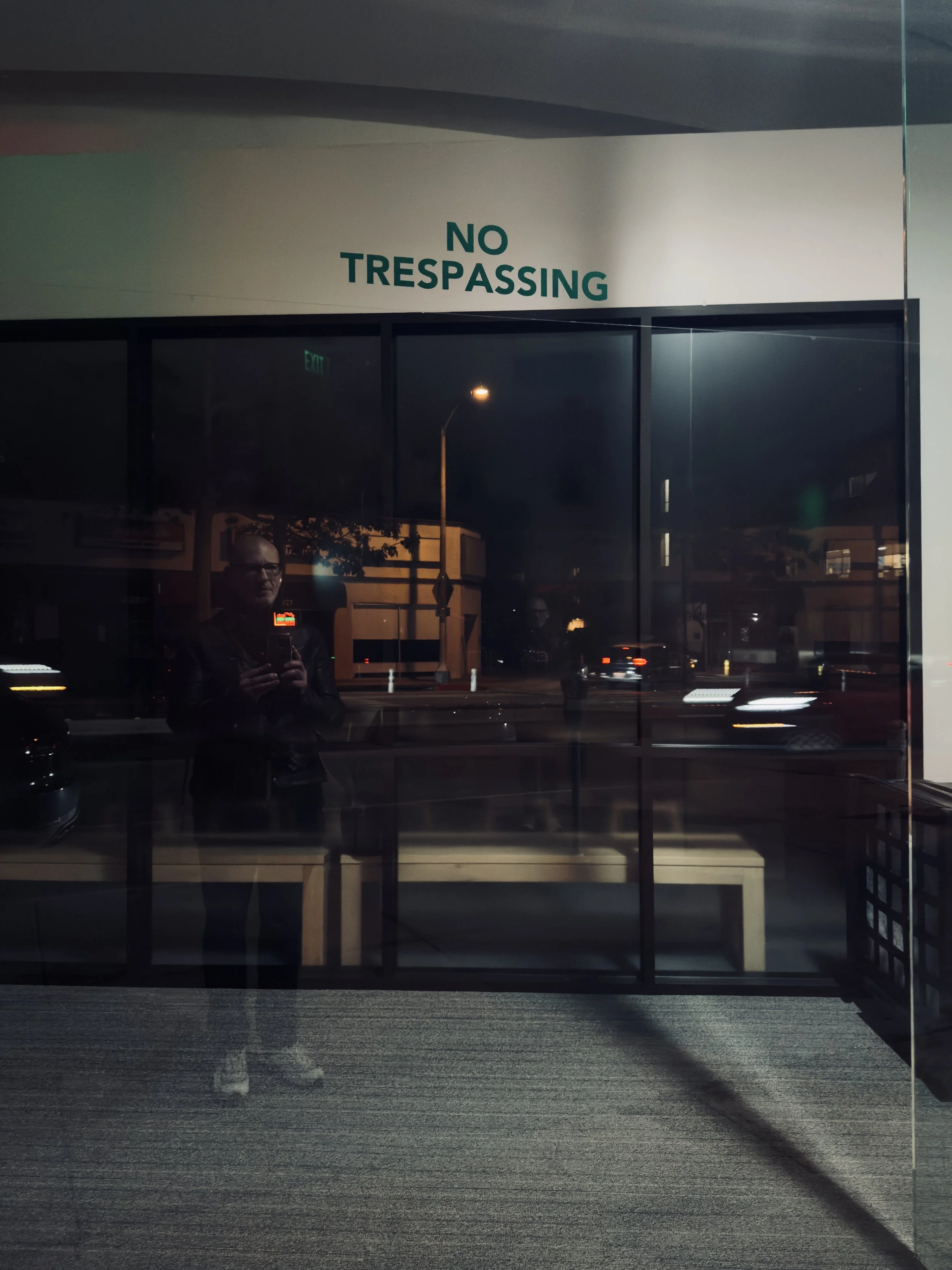 A glass window with a green 'NO TRESPASSING' sign on it, reflecting the street outside at night with cars and streetlights.