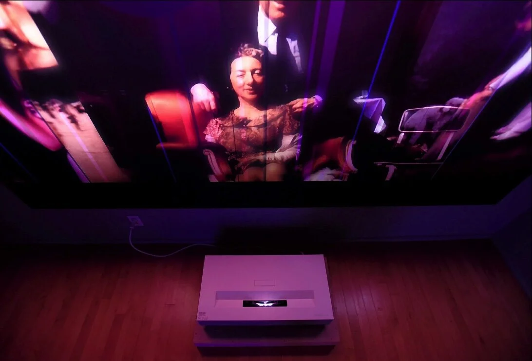 Digital projection on a wall showing an image of a woman seated, with a projector on the floor below.