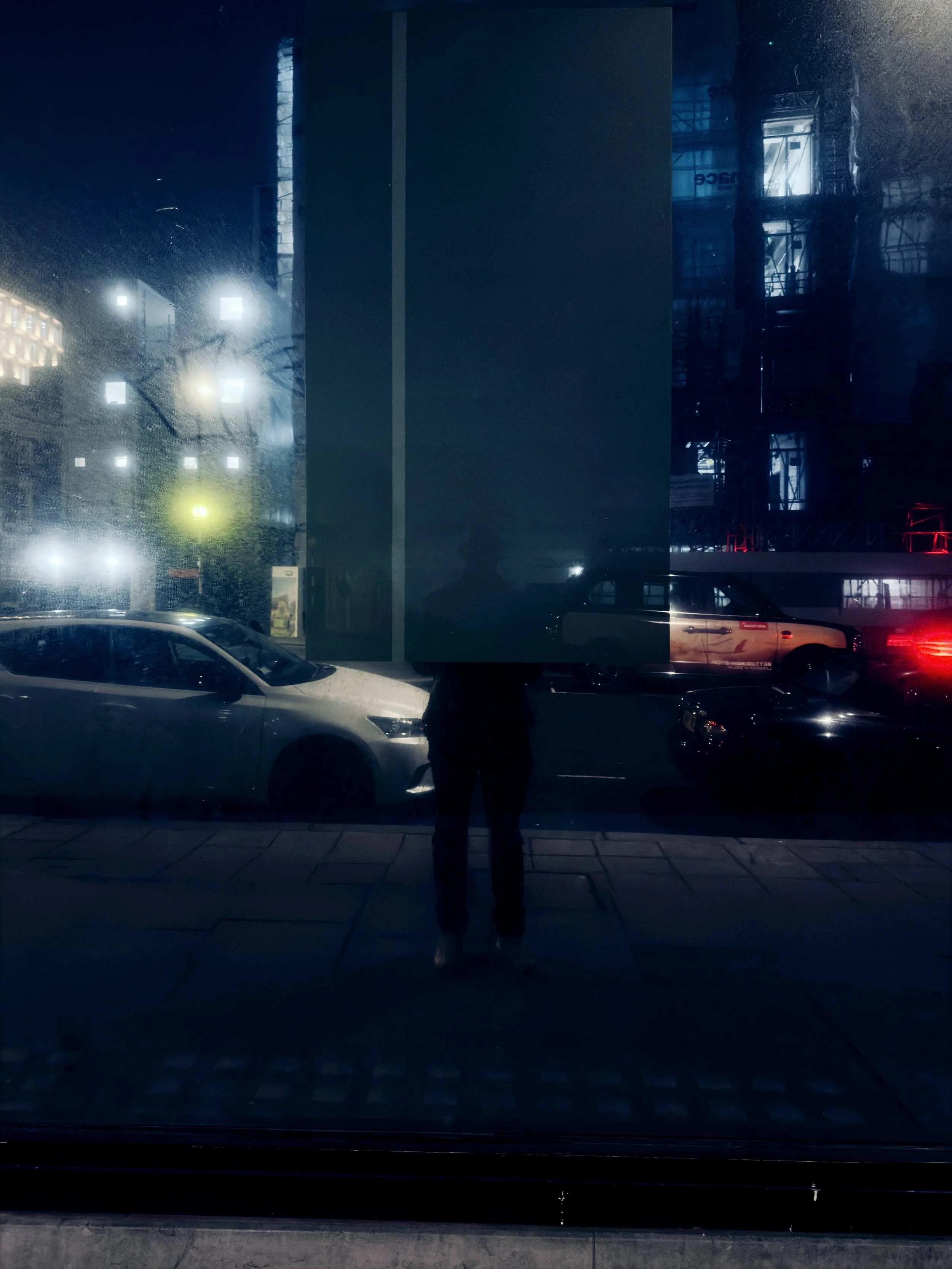 City street scene at night viewed through a large glass window. There are parked cars and reflections of city lights and buildings, with a person standing in the reflection.