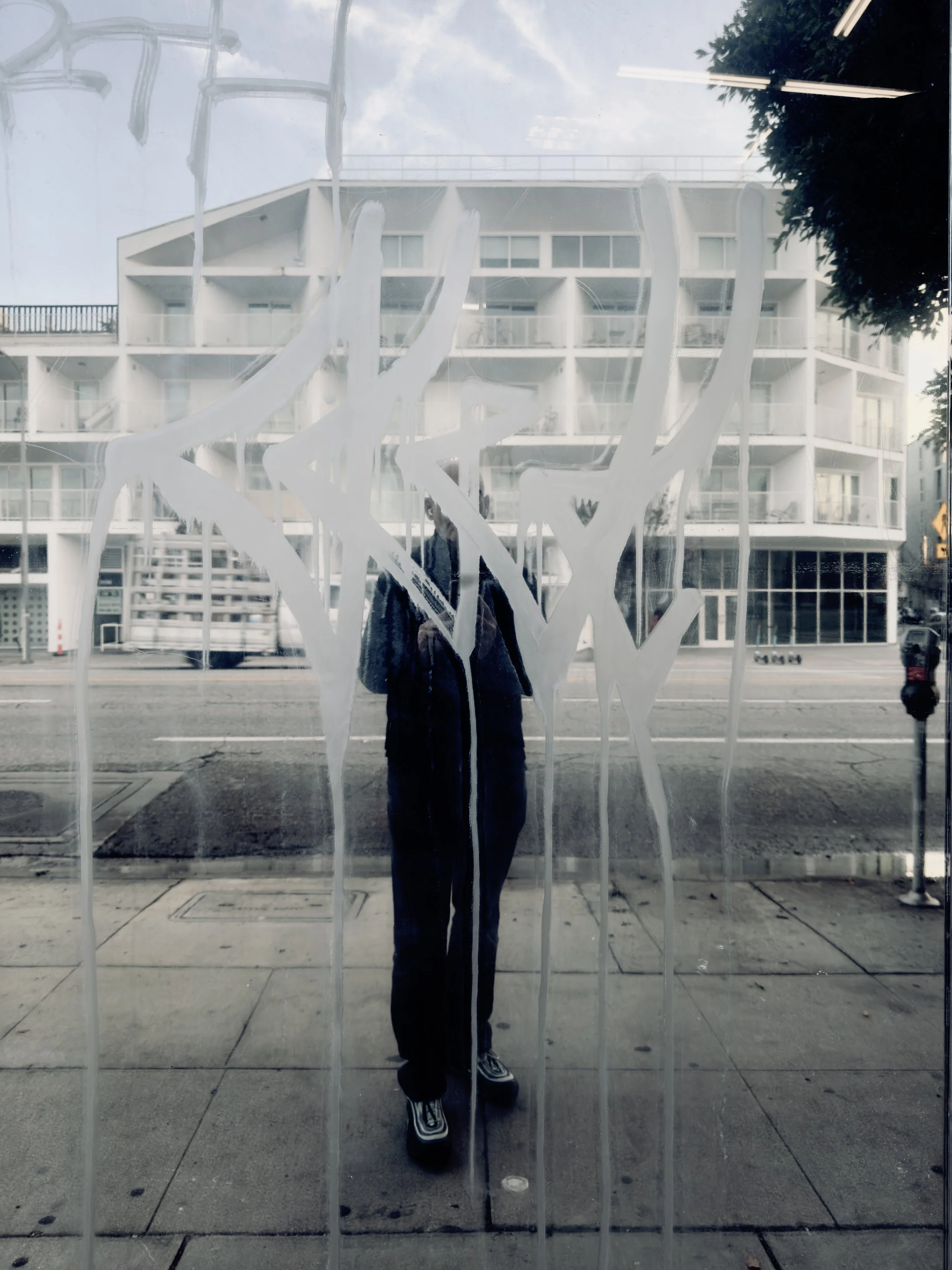 Reflection of a person taking a photo in a glass window with white abstract painted design, street and modern buildings in the background.