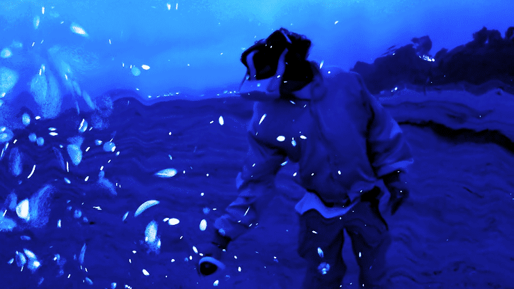 A person in a blue-toned abstract image, possibly distorted by reflections or water, wearing a bulky coat and reaching out.