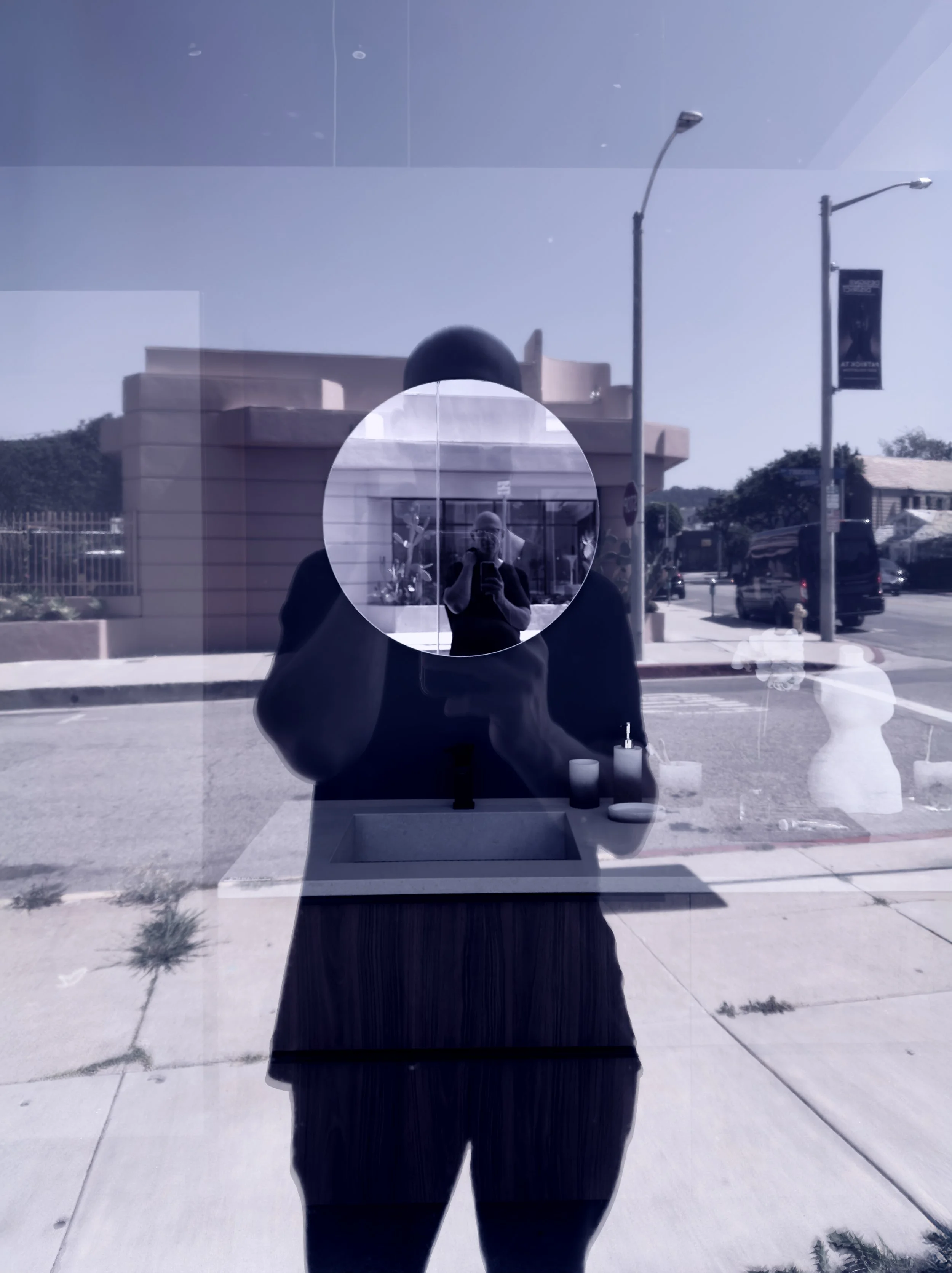 Reflection of a man taking a photo of himself in a glass window, with buildings, streetlights, and cars outside, and bathroom items like a sink and soap dispenser inside.