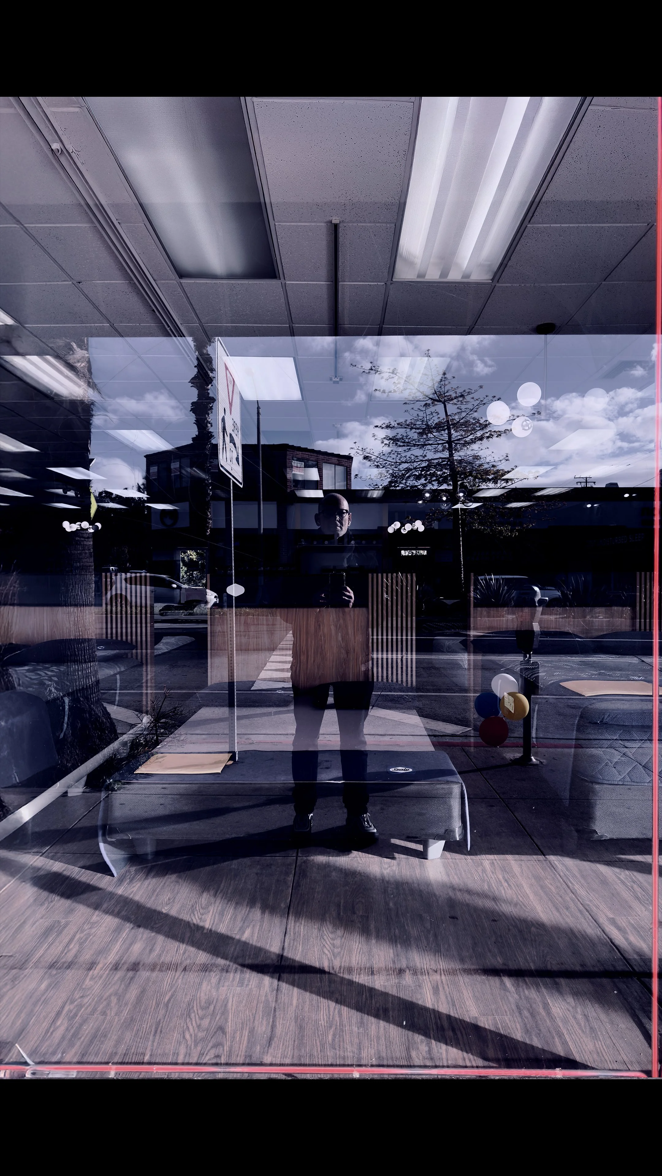 A reflection of a man in a glass window showing the interior ceiling lights and outdoor sky, with trees and buildings outside.