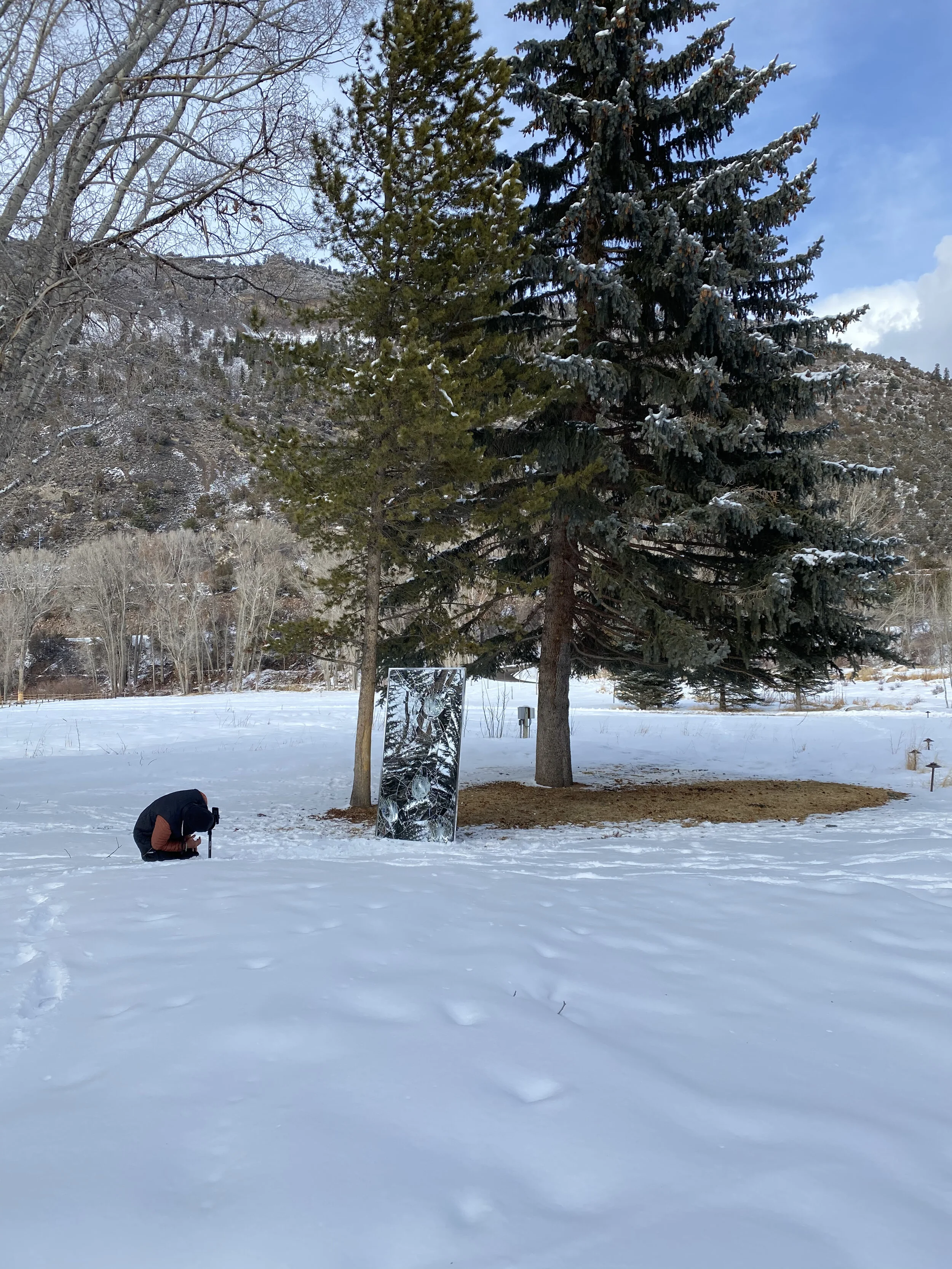 A snowy landscape with a person crouching, taking a photo of a large mirror propped against pine trees. The mirror reflects the snowy and mountainous background.