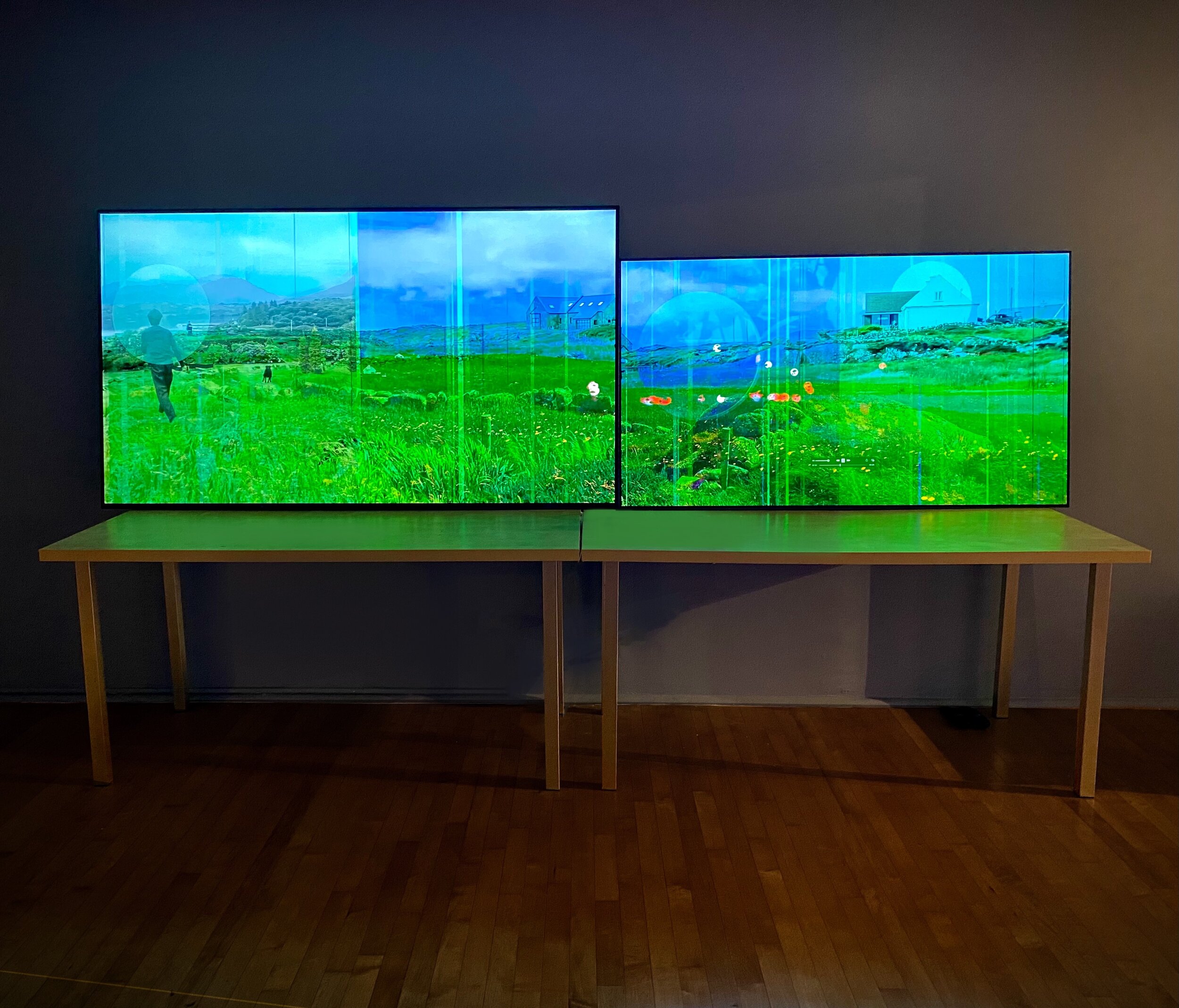 Two large screens displaying a green landscape with a figure and buildings, mounted on a wooden table against a dark wall.