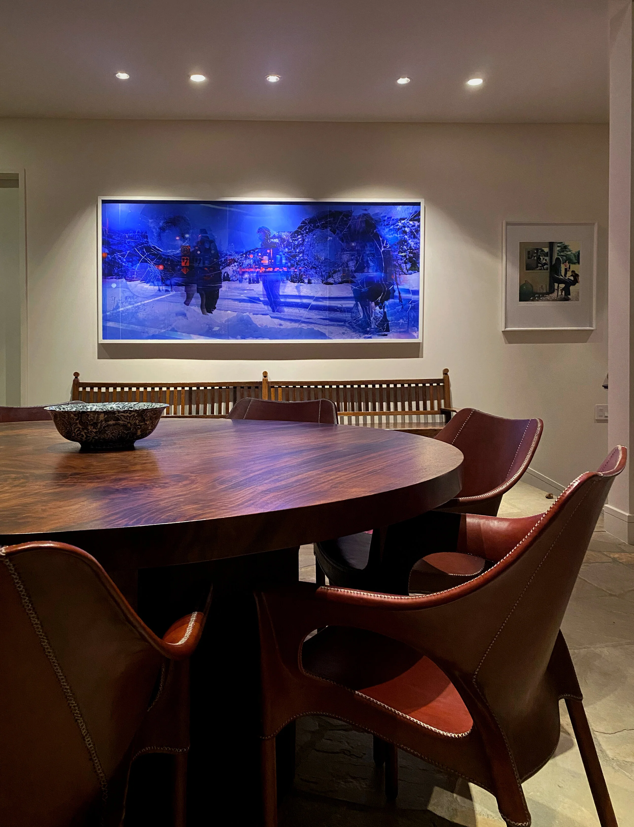 Dining room with a round wooden table, leather chairs, a decorative bowl, large blue-themed artwork on the wall, small framed picture, and overhead lighting.