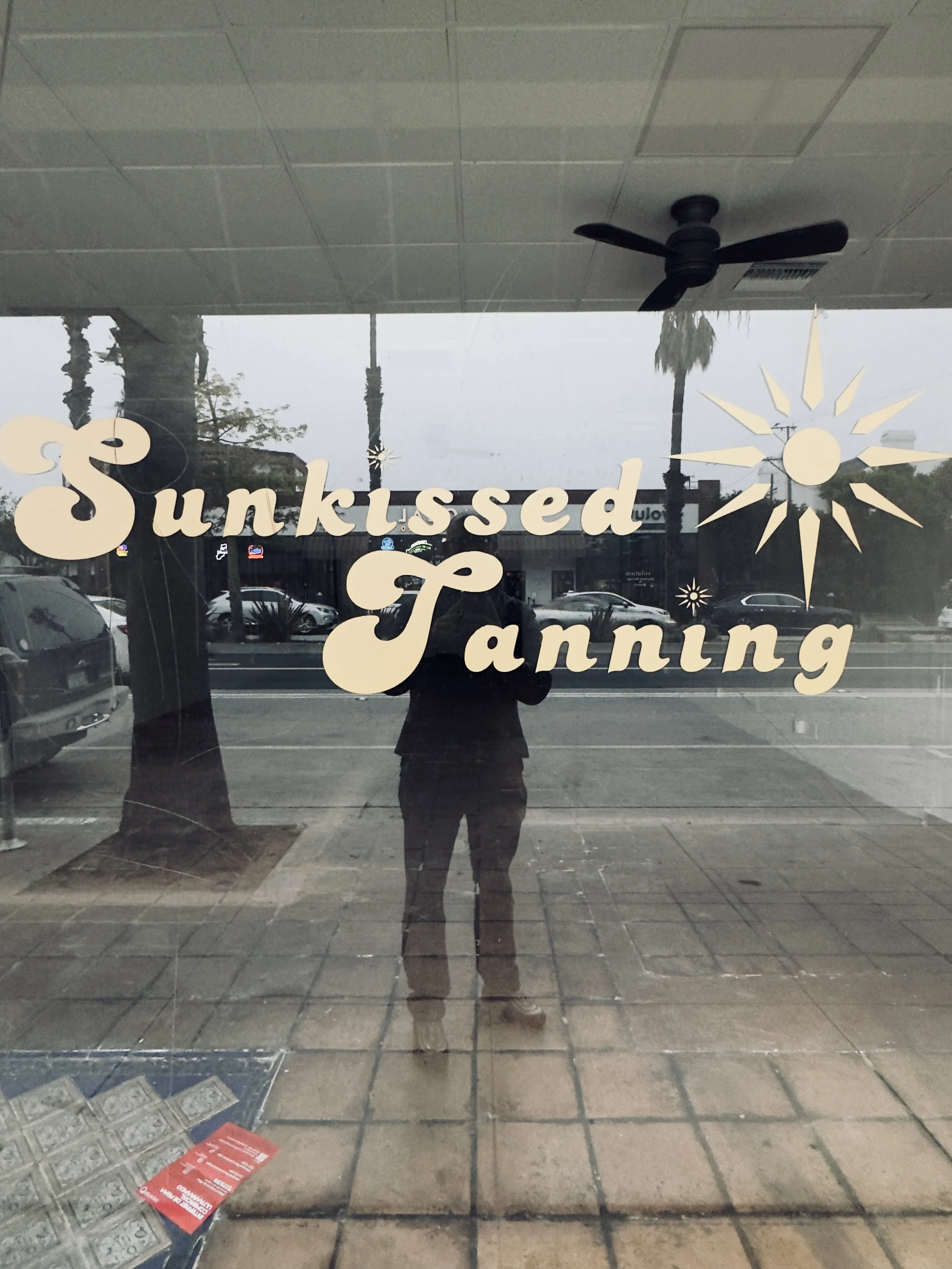 Storefront window with the name "Sunkissed Tanning" and a sun graphic. The reflection of a person taking a photo is visible, along with the street and parked cars outside.