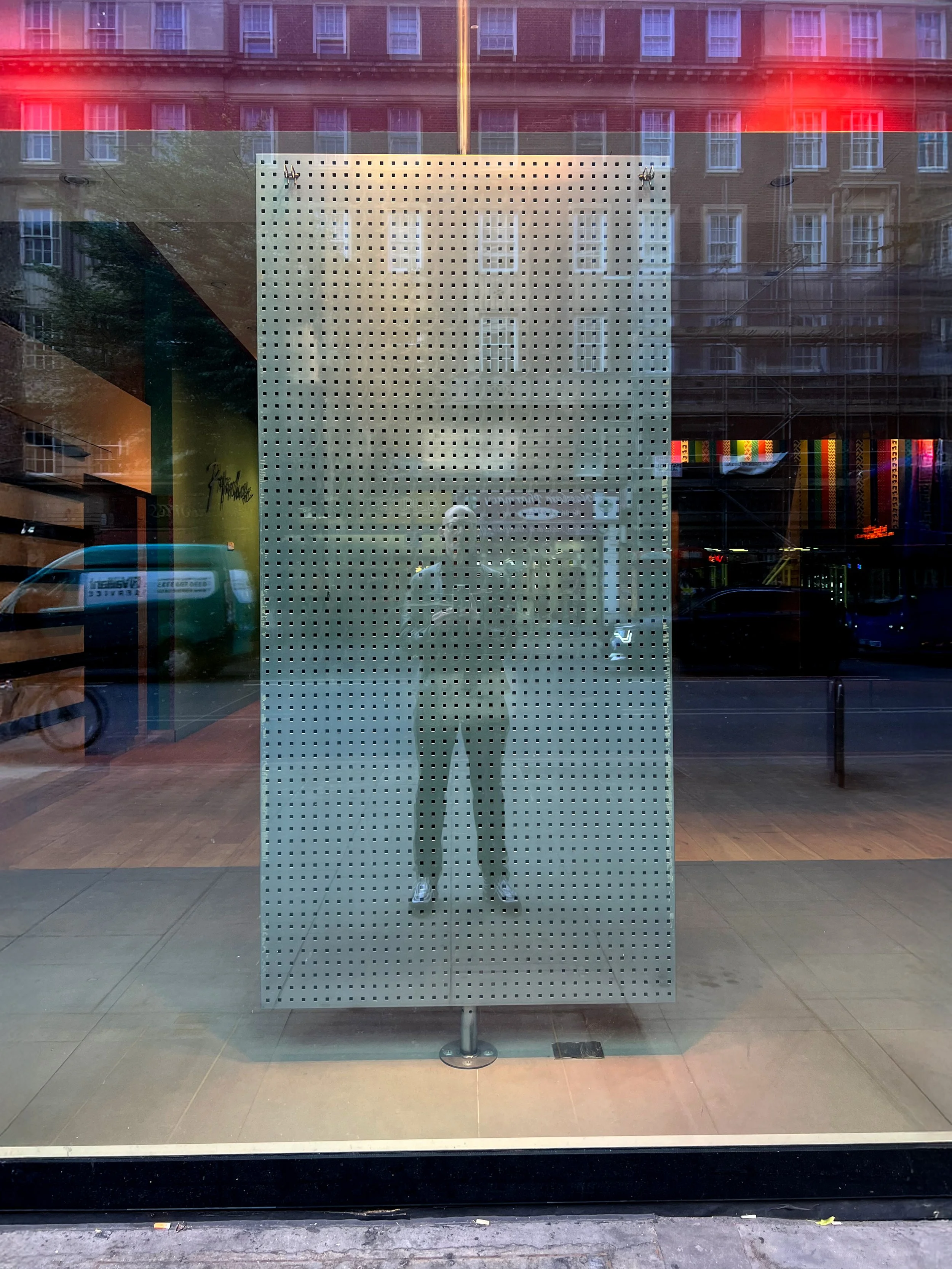 Reflection of a person taking a photo in a large window with a perforated metal panel and an urban street scene in the background.
