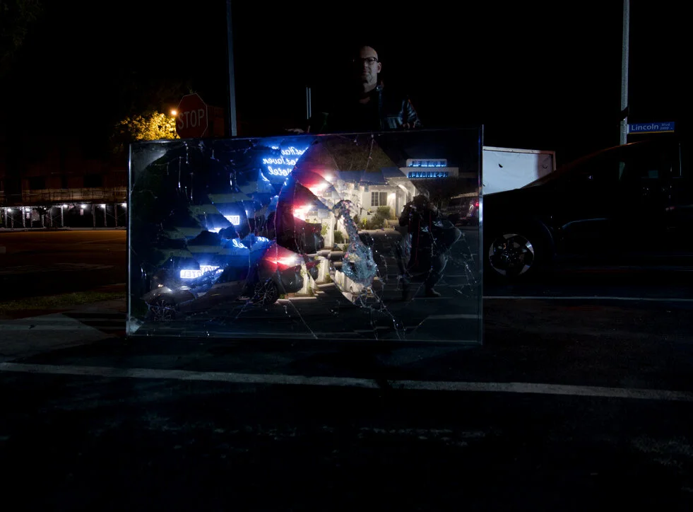 Broken mirror reflecting neon lights and a man at night, parking lot scene.
