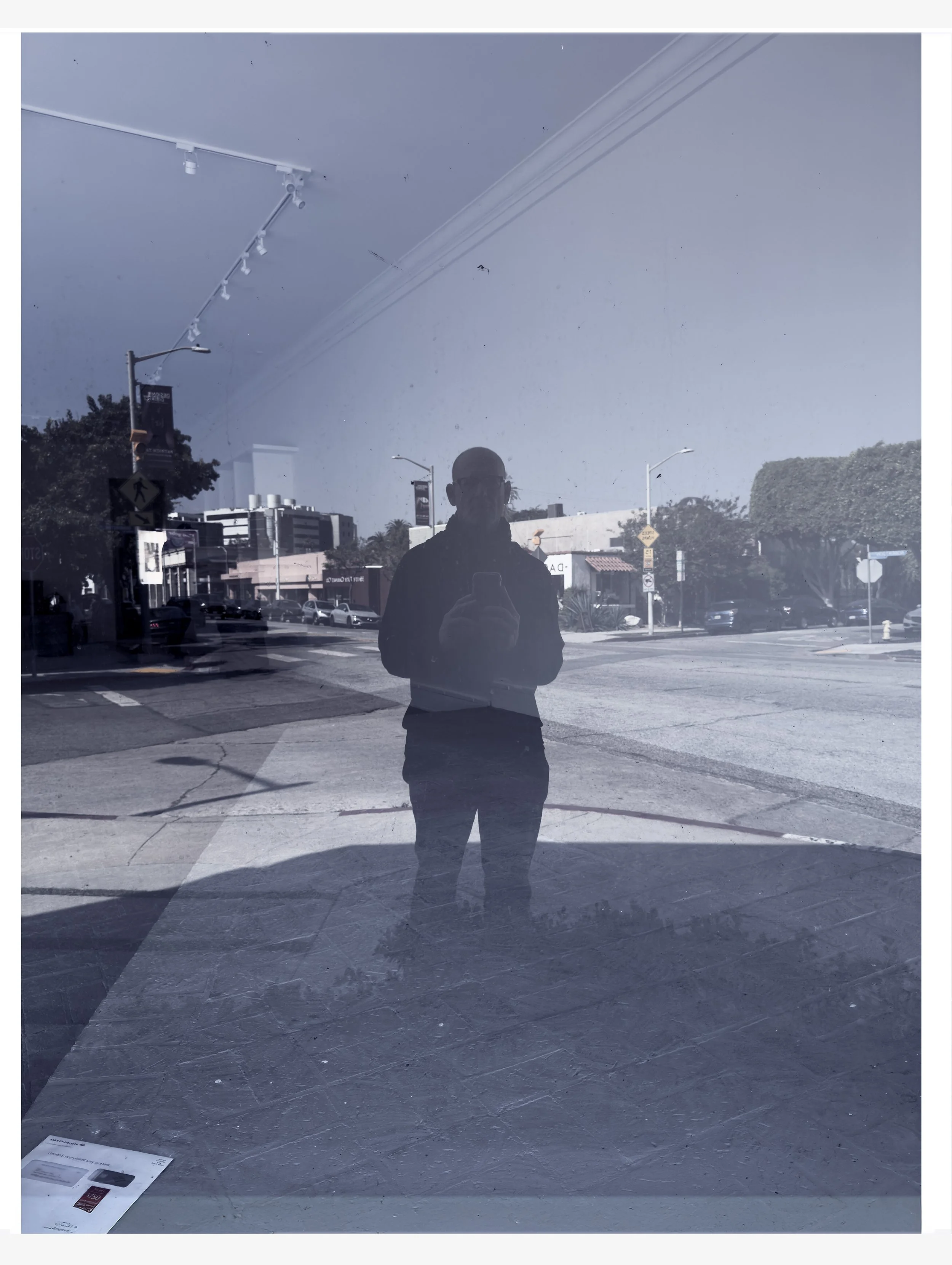 Photograph of a street scene reflected in a glass window, with a silhouette of a person taking a photo of the reflection, and urban buildings, street signs, and trees visible outside.